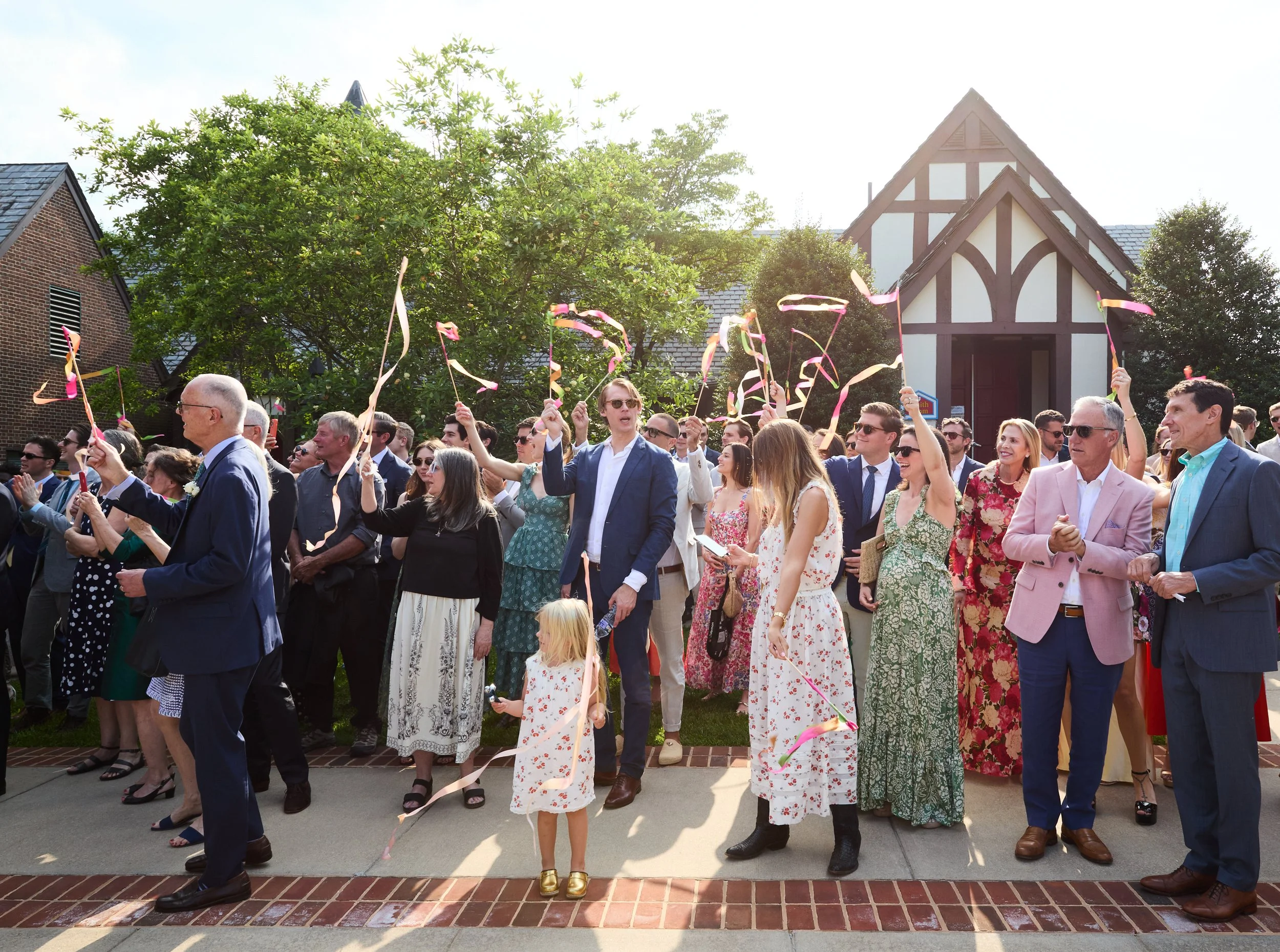 A group of people celebrating outdoors with streamers and confetti, dressed in colorful and formal attire, in front of a Tudor-style building and trees.