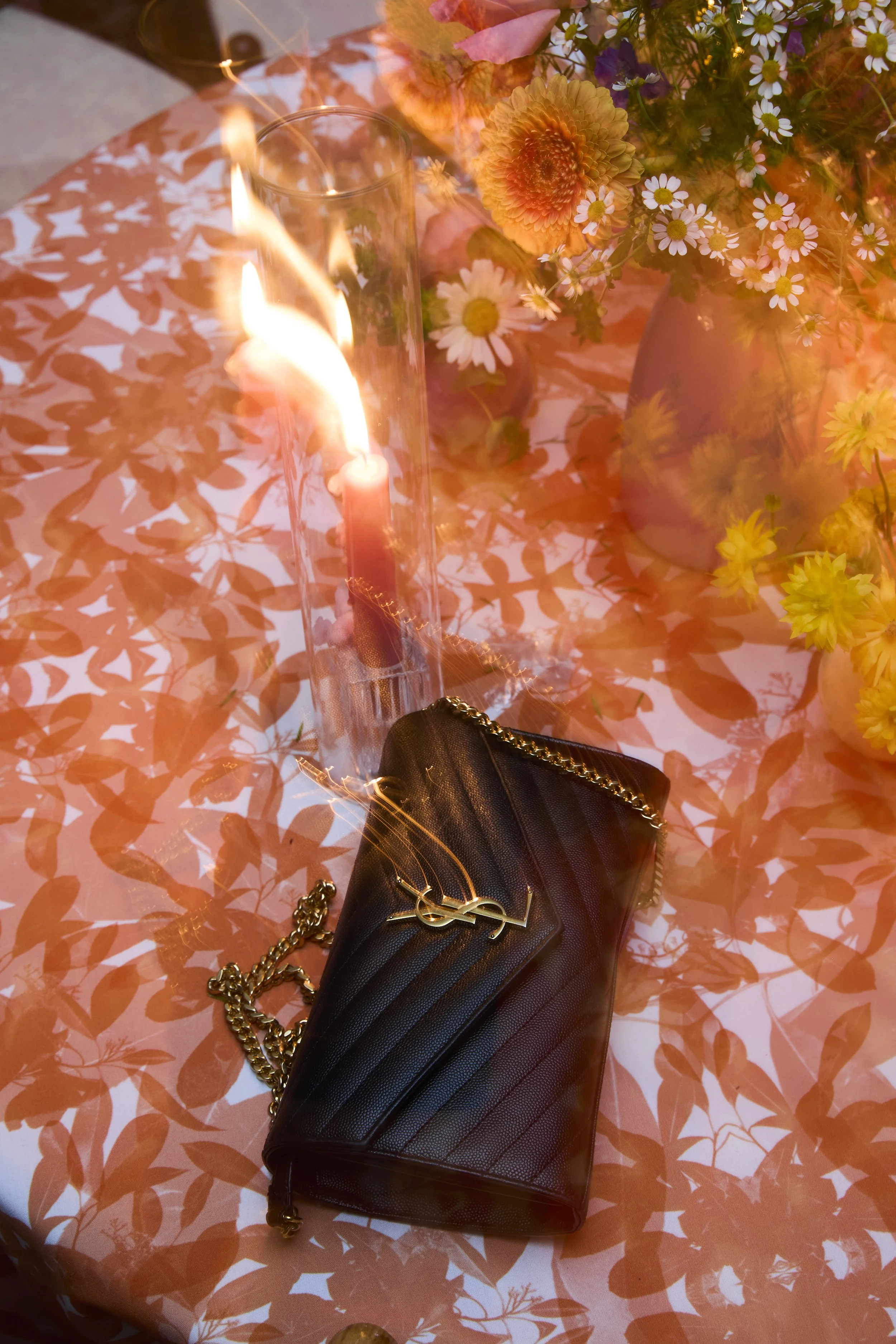 A table setting with a lit candle inside a glass holder, a black Yves Saint Laurent purse with a gold chain and logo, and a bouquet of colorful flowers including daisies in vases. The table has a patterned tablecloth with orange and white floral desi