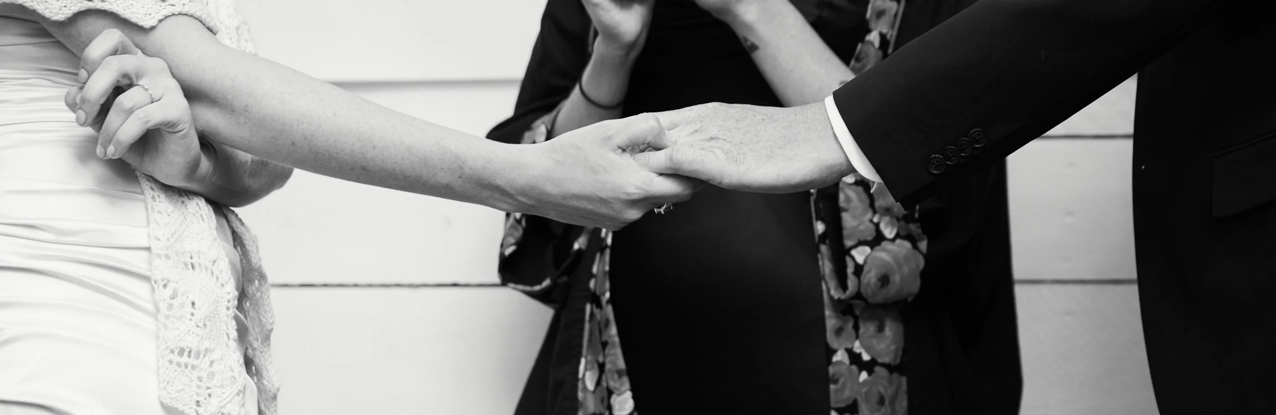 A black and white photograph of a wedding ceremony with a bride and groom shaking hands, the bride's arm and hand are visible, and the groom is wearing a suit.