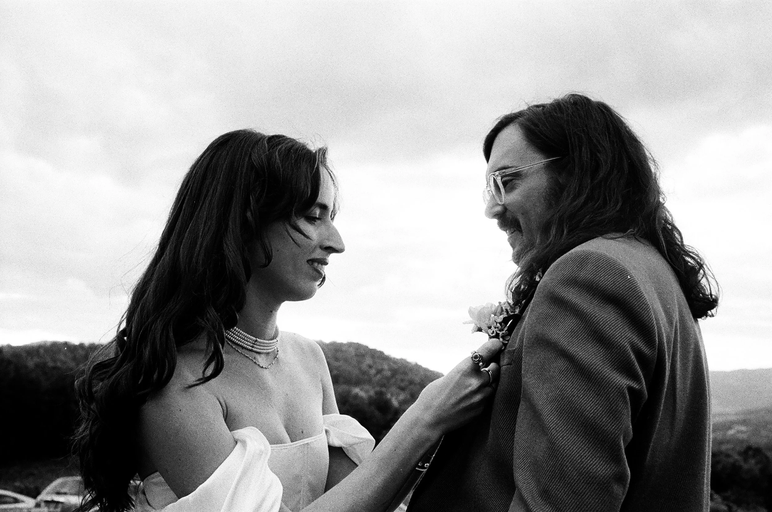 A black-and-white photo of a smiling woman with long dark hair and a woman with long wavy hair and glasses, dressed in a suit, sharing a tender moment outdoors