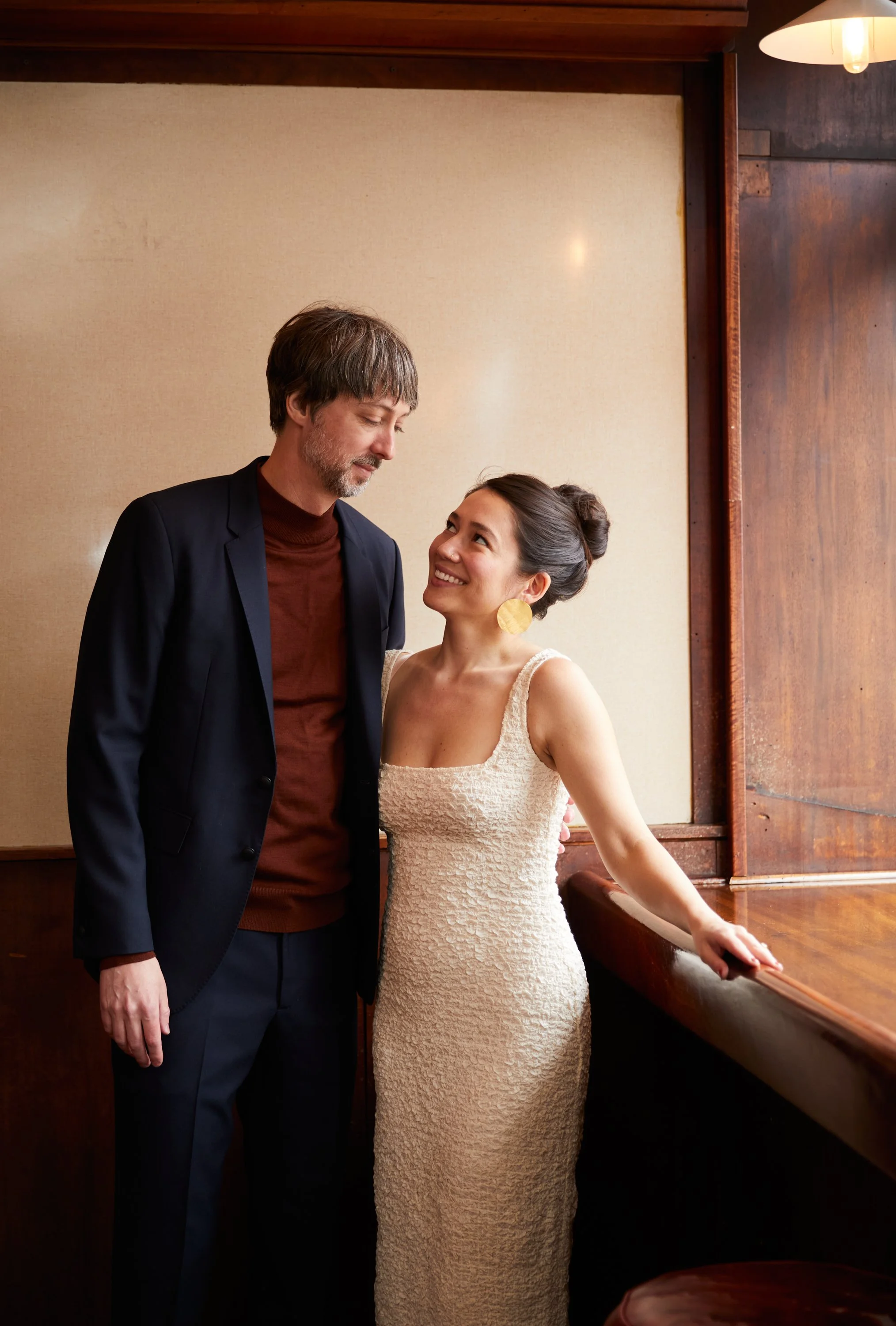 A man and woman smiling and looking at each other indoors, with the woman leaning on a wooden bar or counter.