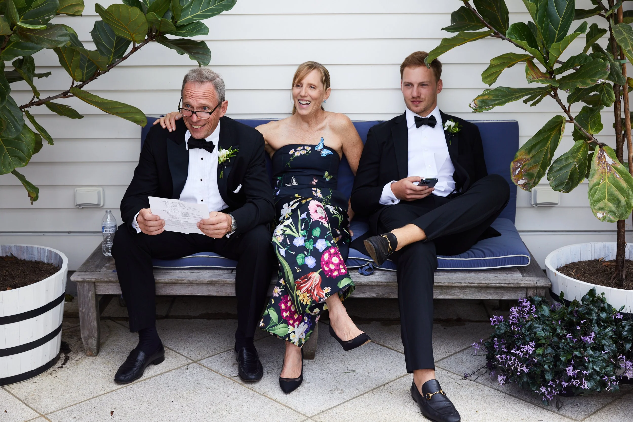Three people dressed in formal attire sitting on a wooden bench with pillows outdoors, two men in tuxedos and a woman in a floral dress, surrounded by potted plants.