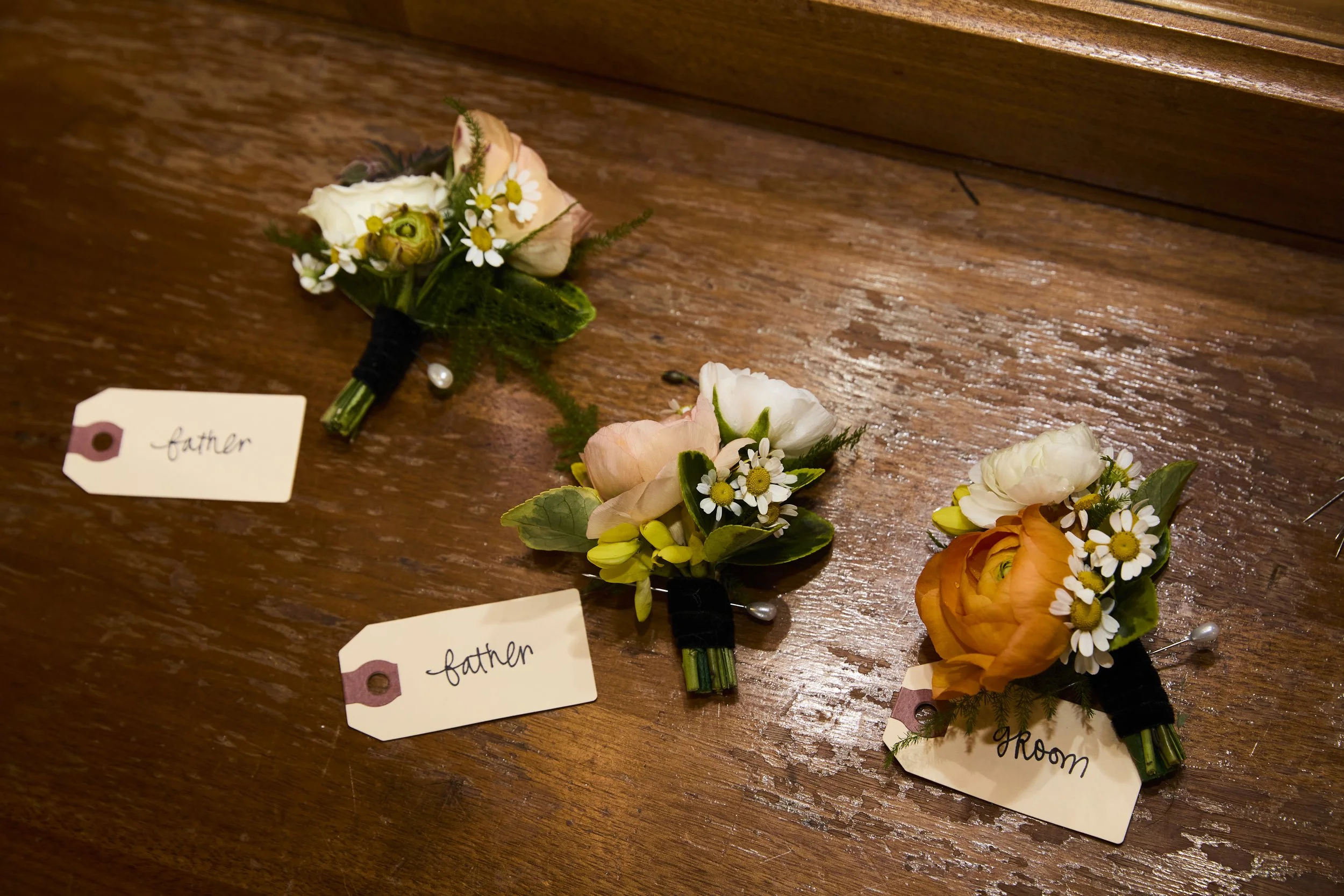 Three floral boutonnieres with tags reading 'father' and 'room' laid on a wooden surface.