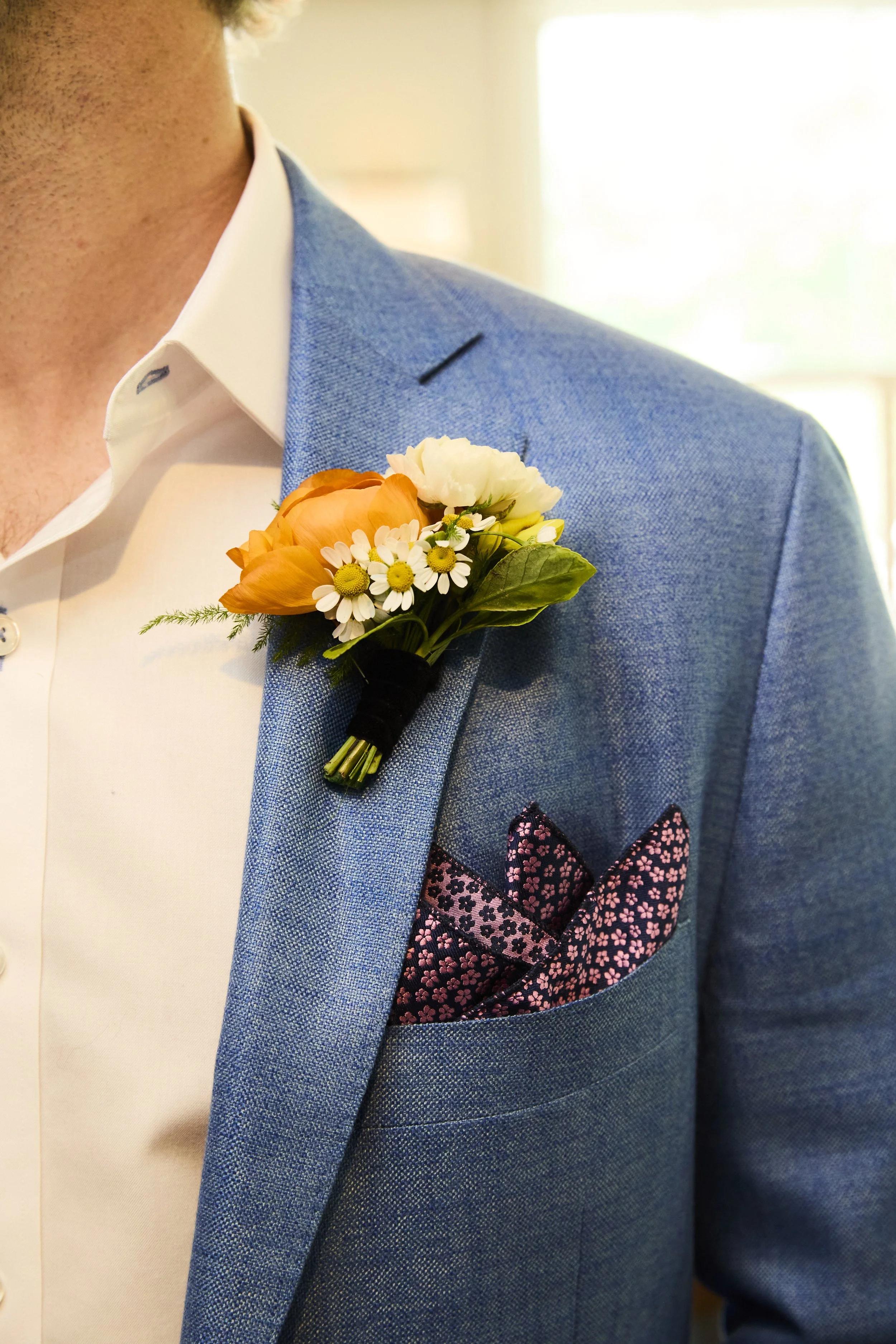 Close-up of a man wearing a light blue suit jacket, white dress shirt, and a floral pocket square. He has a boutonniere with orange and white flowers on his lapel.