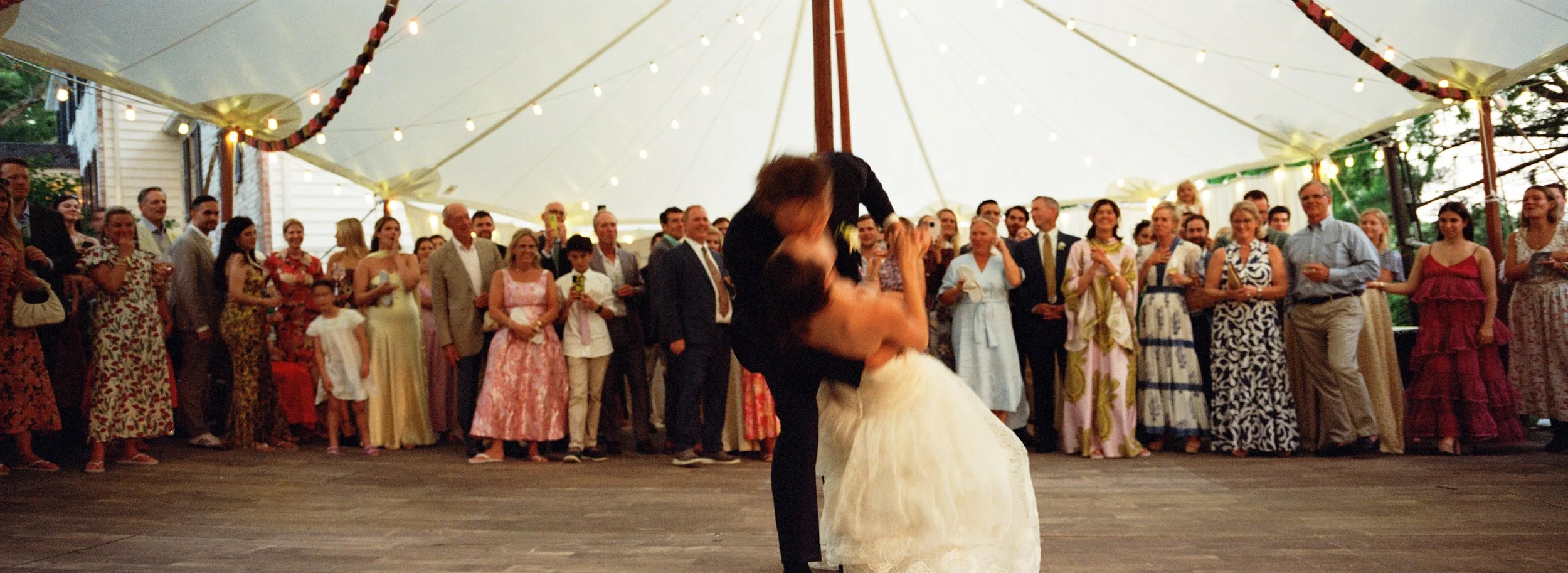 A wedding dance under a large tent decorated with string lights, with a bride and groom dancing while surrounded by friends and family in formal and semi-formal attire.