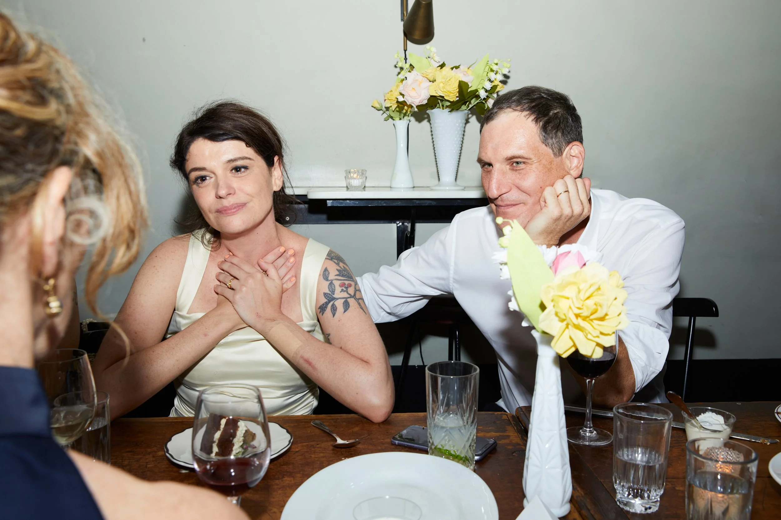 A man and a woman sit at a dinner table with flowers, drinks, and dessert, smiling and holding hands over their hearts in an intimate setting.