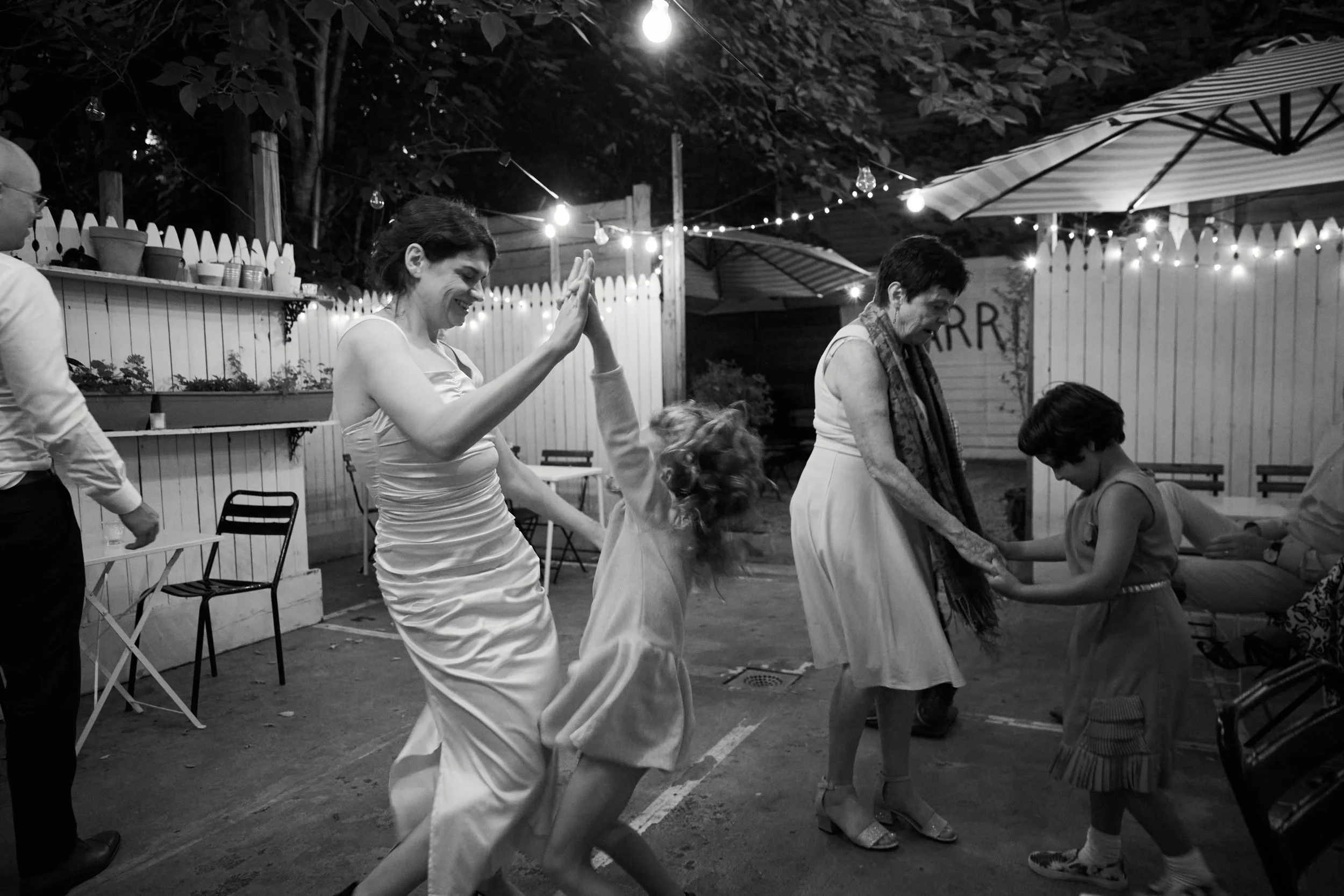 People dancing and enjoying at an outdoor celebration or party, with string lights and a white picket fence in the background.