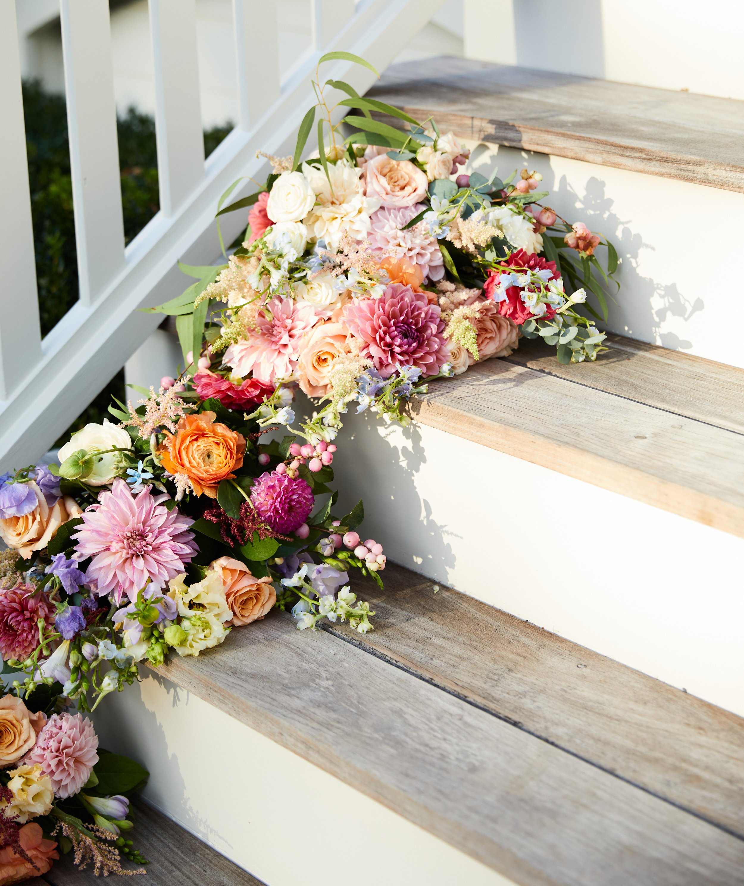 A colorful flower arrangement with various pink, peach, white, and purple flowers draped over outdoor wooden steps at a wedding or event.