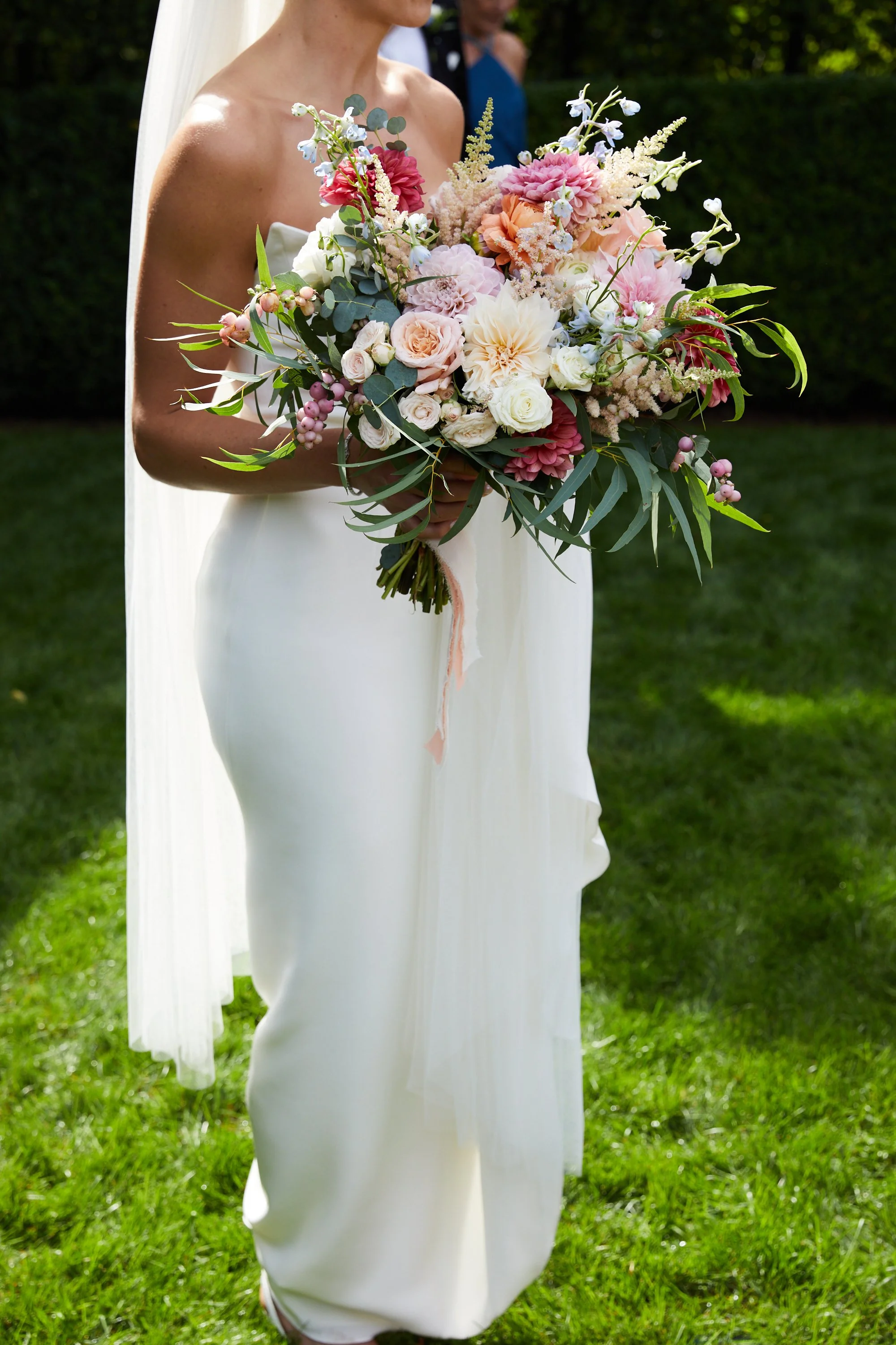 A bride holding a large bouquet of pink, white, and pastel flowers outdoors on a green lawn.