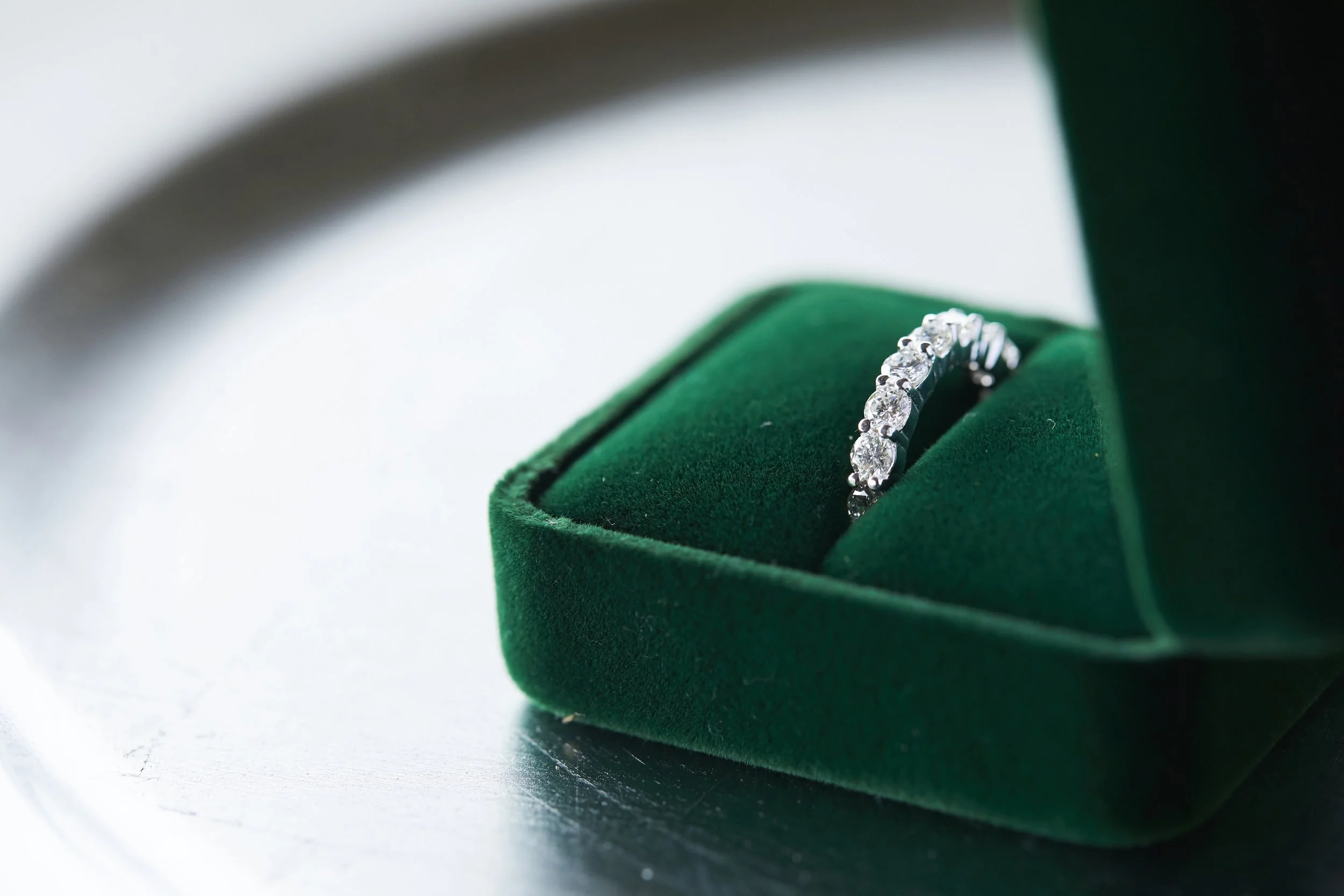 A diamond engagement ring in a green velvet ring box on a silver surface.