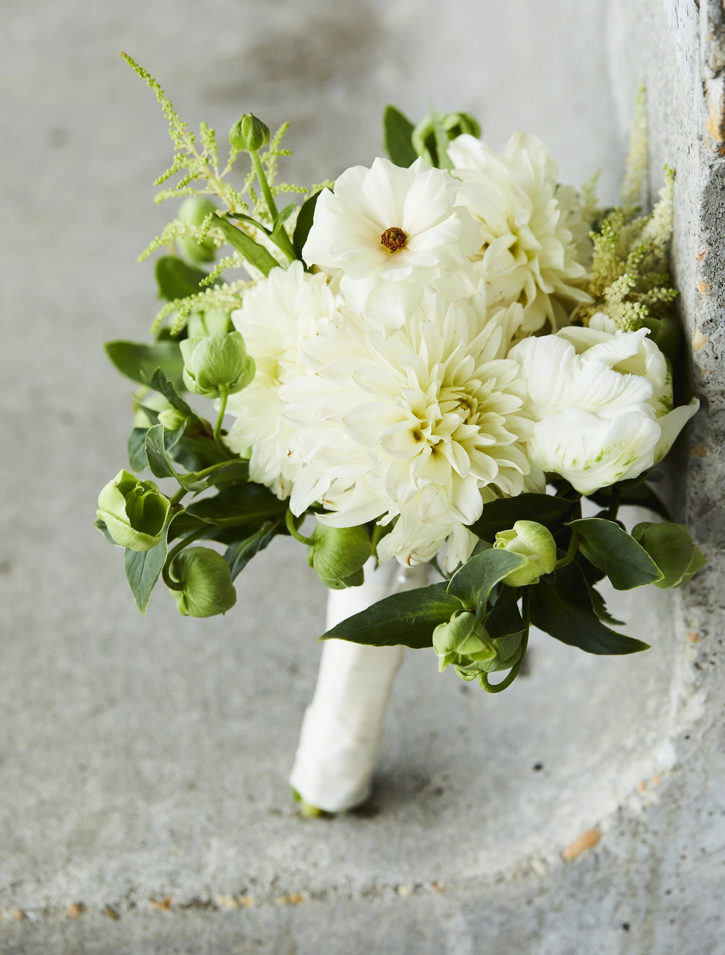 Bouquet of white flowers including dahlias and roses with green leaves, placed on a concrete surface.