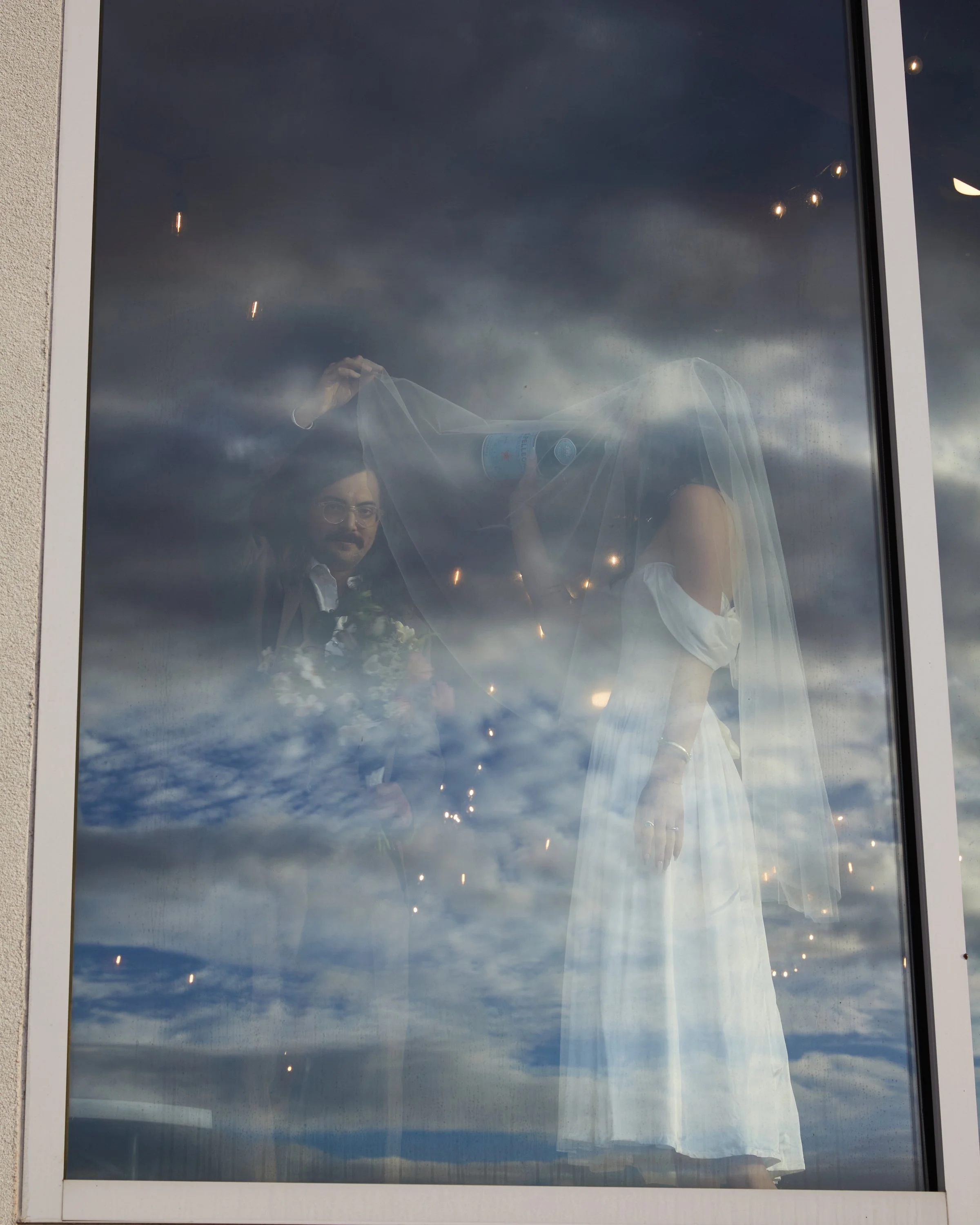 Reflection of a bride and groom inside a window, with the sky and clouds visible outside, creating a layered view.