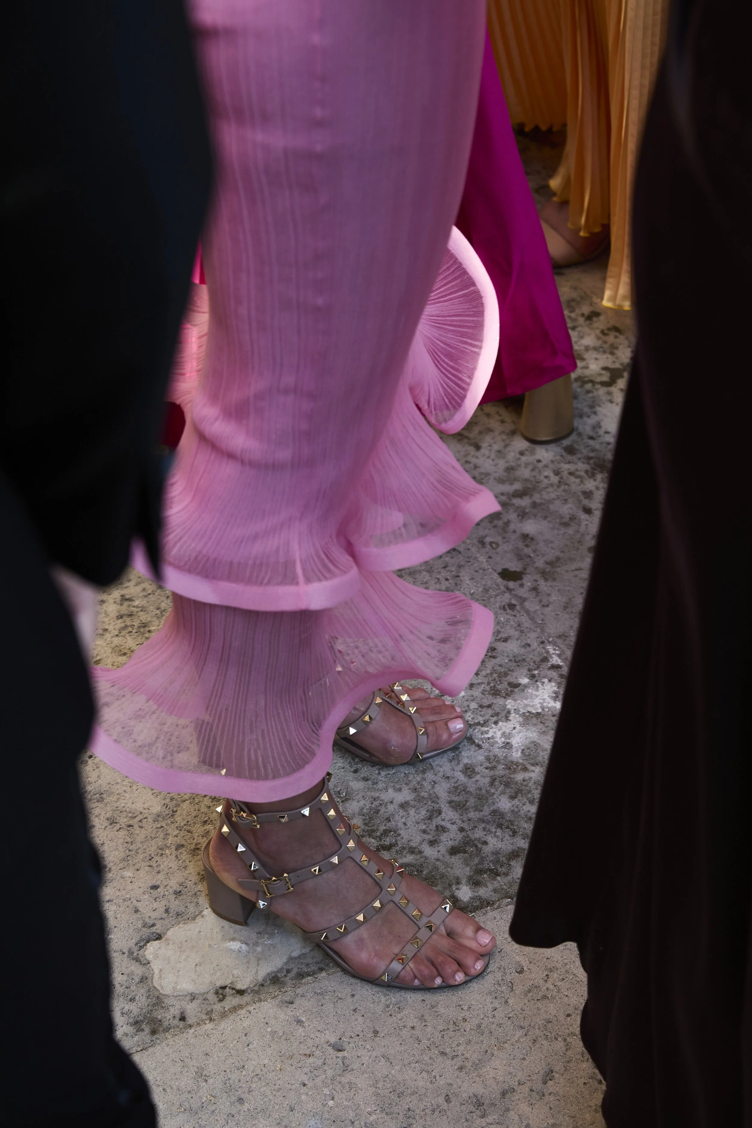 Close-up of a woman's legs wearing a pink, ruffled dress and embellished high-heeled sandals, seen from among seated and standing people at an event.