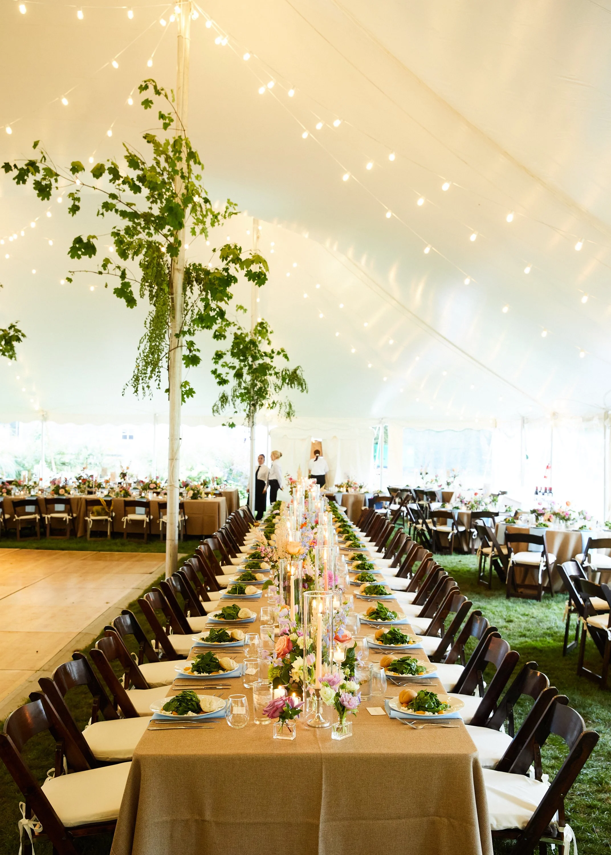 Long banquet table adorned with flowers, candles, and place settings under a tent with string lights and greenery.