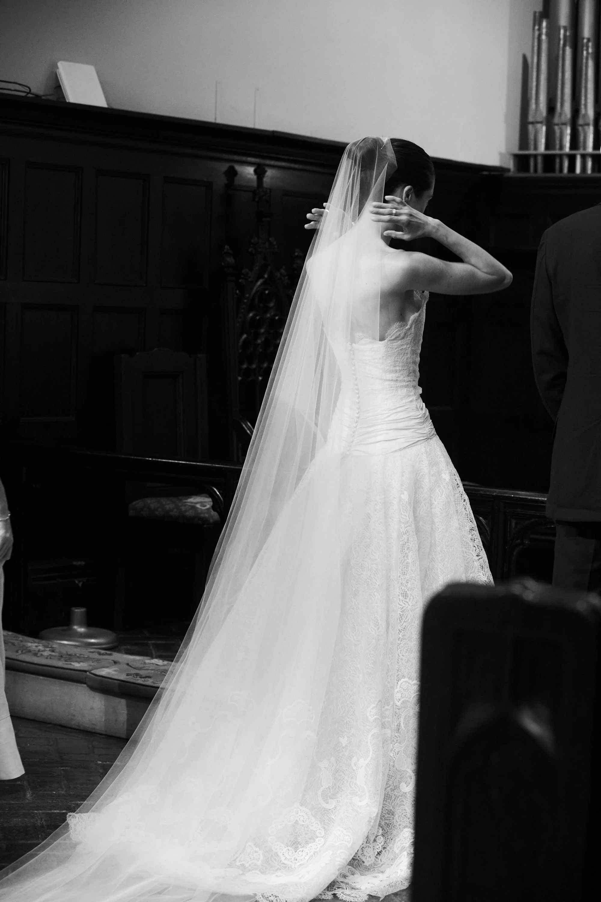 A bride in a wedding dress with a long veil adjusting her hair in a room with wooden paneling and a bookshelf.