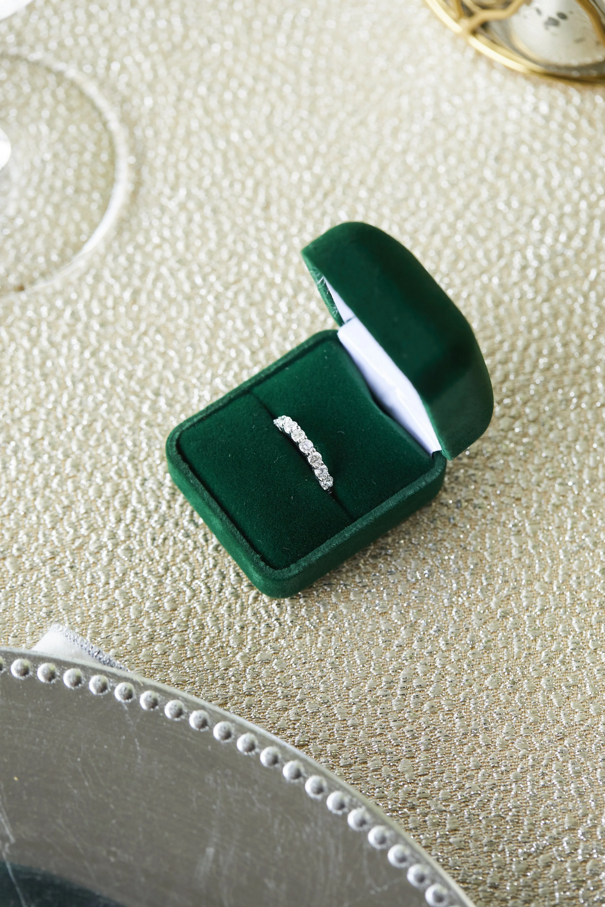 A diamond ring in a green velvet jewelry box on a glittery gold surface.