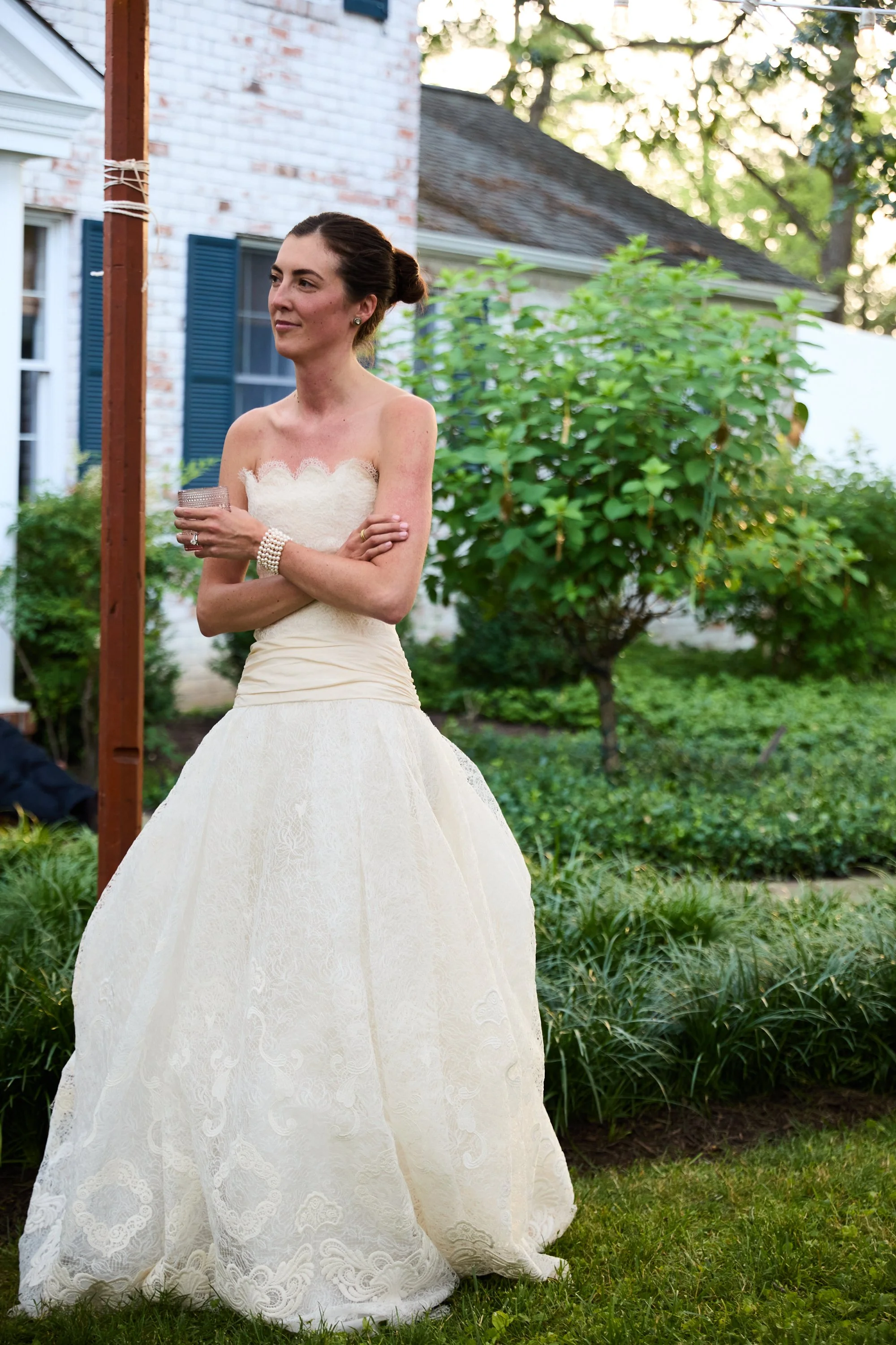 A woman in a white strapless wedding dress stands outdoors in a garden, with her arms crossed and holding a glass, with trees, bushes, a house with blue shutters, and a wooden post in the background
