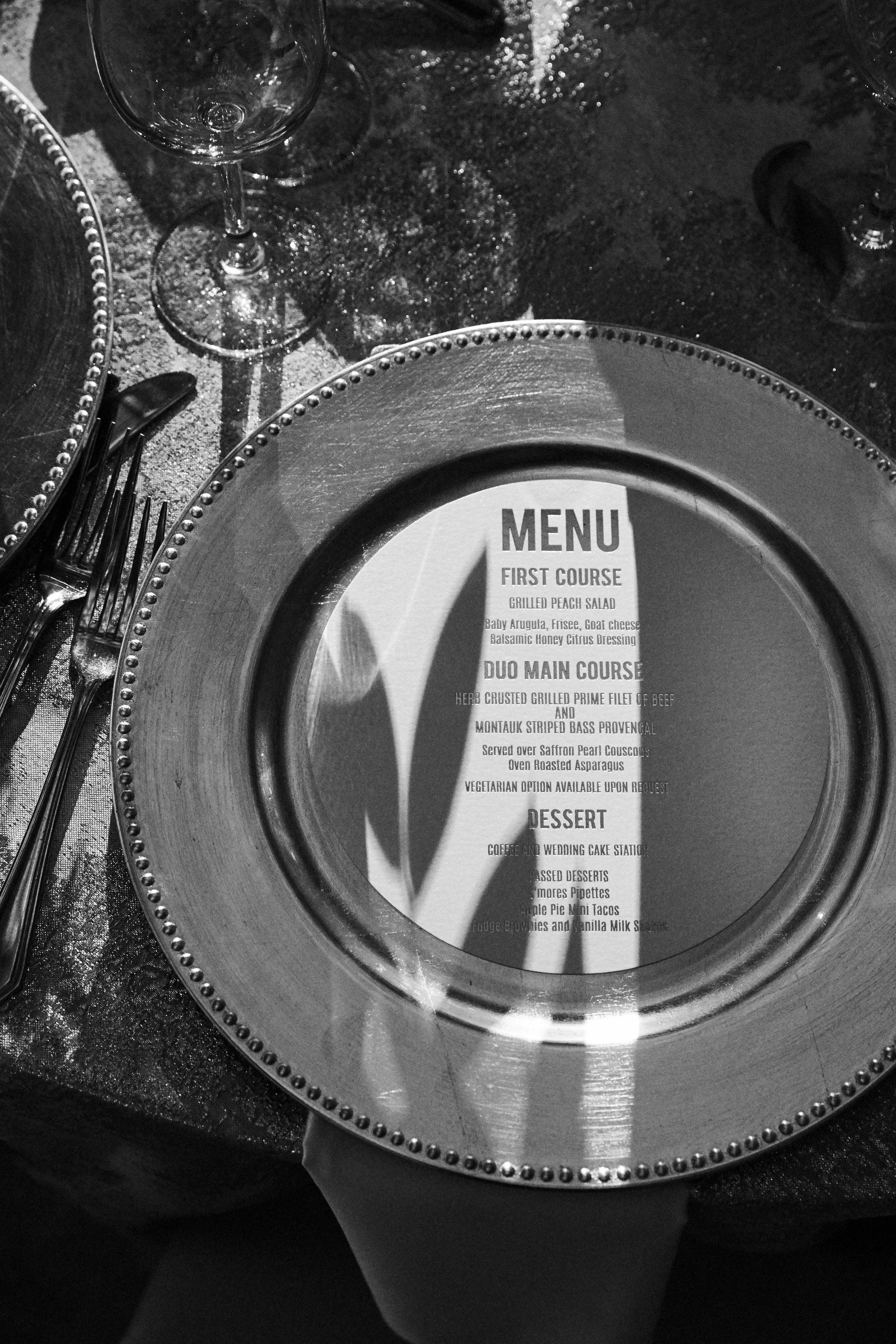 A restaurant table setting with a decorative plate, set of cutlery, wine glasses, and a menu under the plate. The menu lists a three-course meal including a shrimp and goat cheese salad, grilled beef, and various desserts.