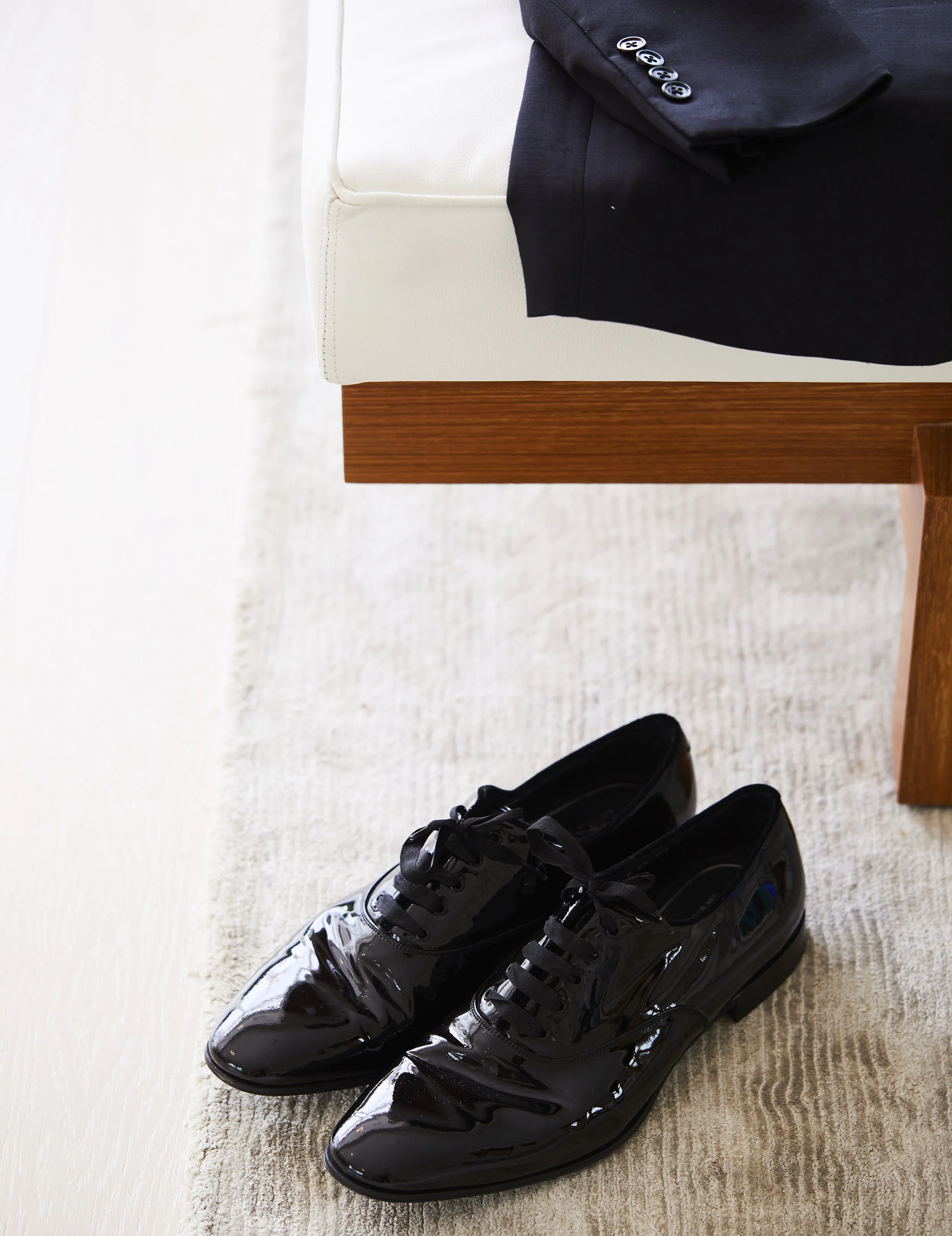 Pair of shiny black dress shoes on a light-colored carpet near a white sofa with dark clothing draped over the side.