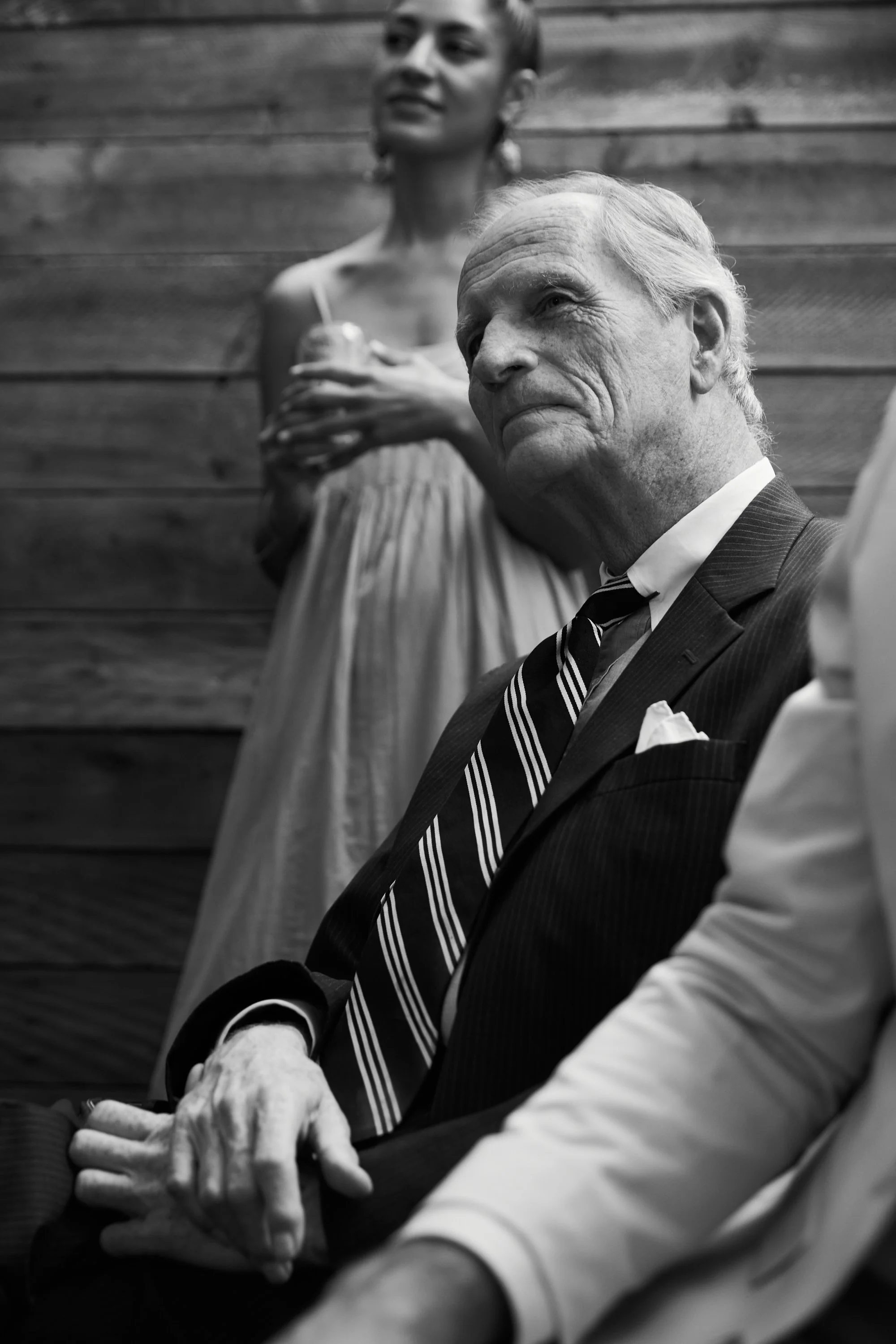 Black and white photo of an elderly man in a suit and striped tie, sitting with his hands clasped, with a woman in a dress standing behind him, holding a drink, against a wooden wall background.
