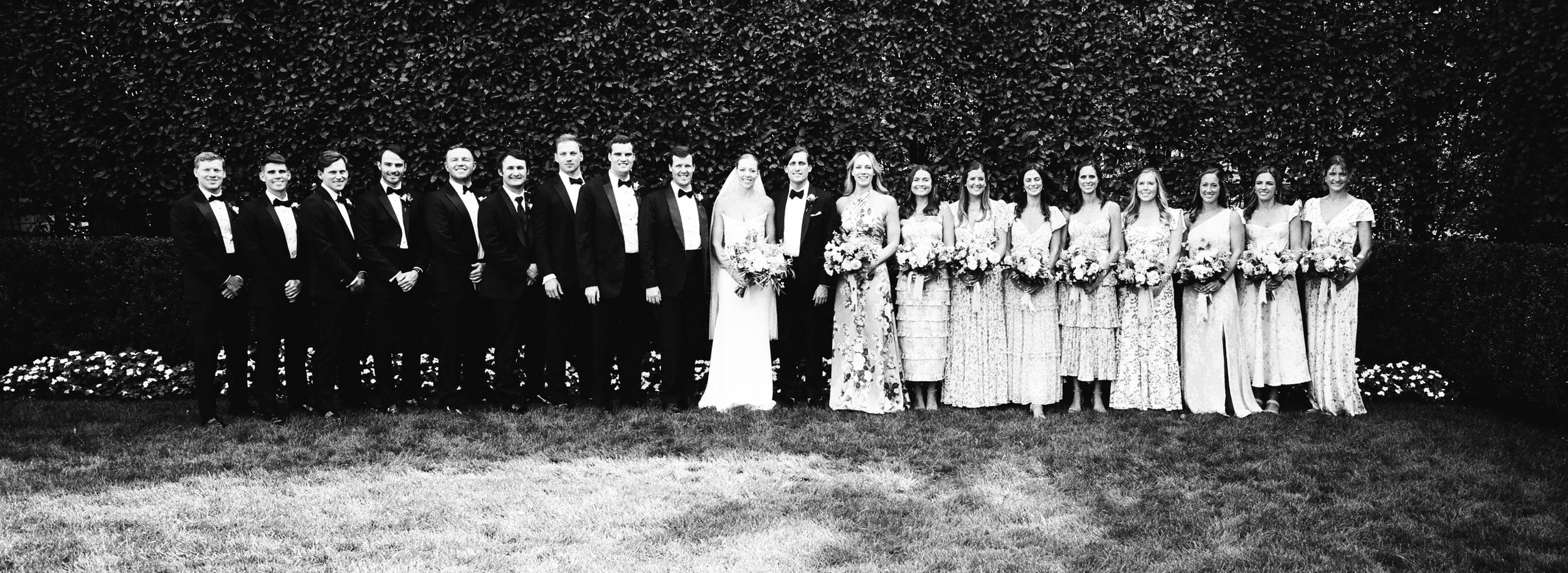 Black and white photo of a wedding party, with men in tuxedos and women in floral dresses, standing outdoors in front of a hedge