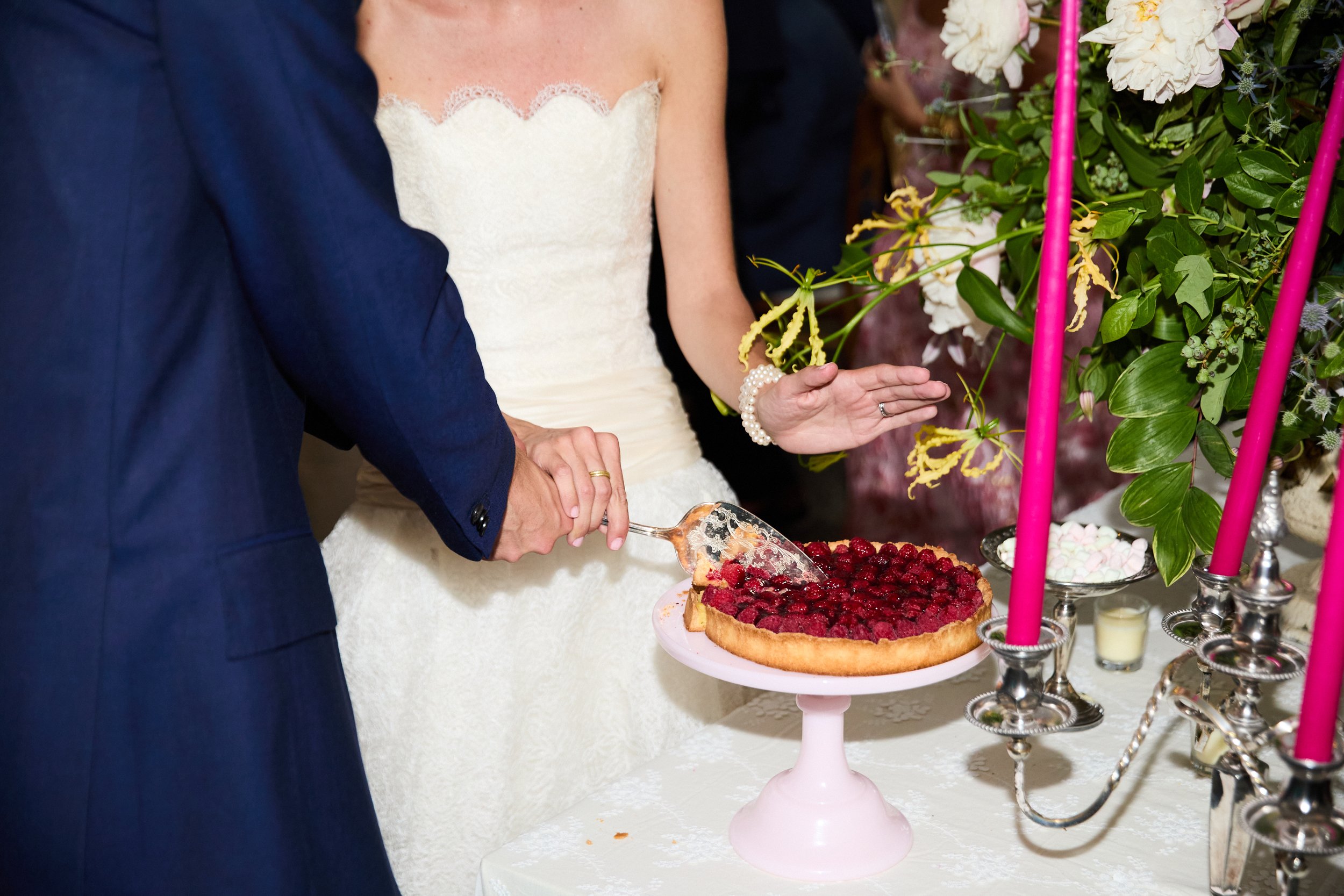 A bride and groom slicing a raspberry tart at their wedding reception, surrounded by flowers and pink candles.