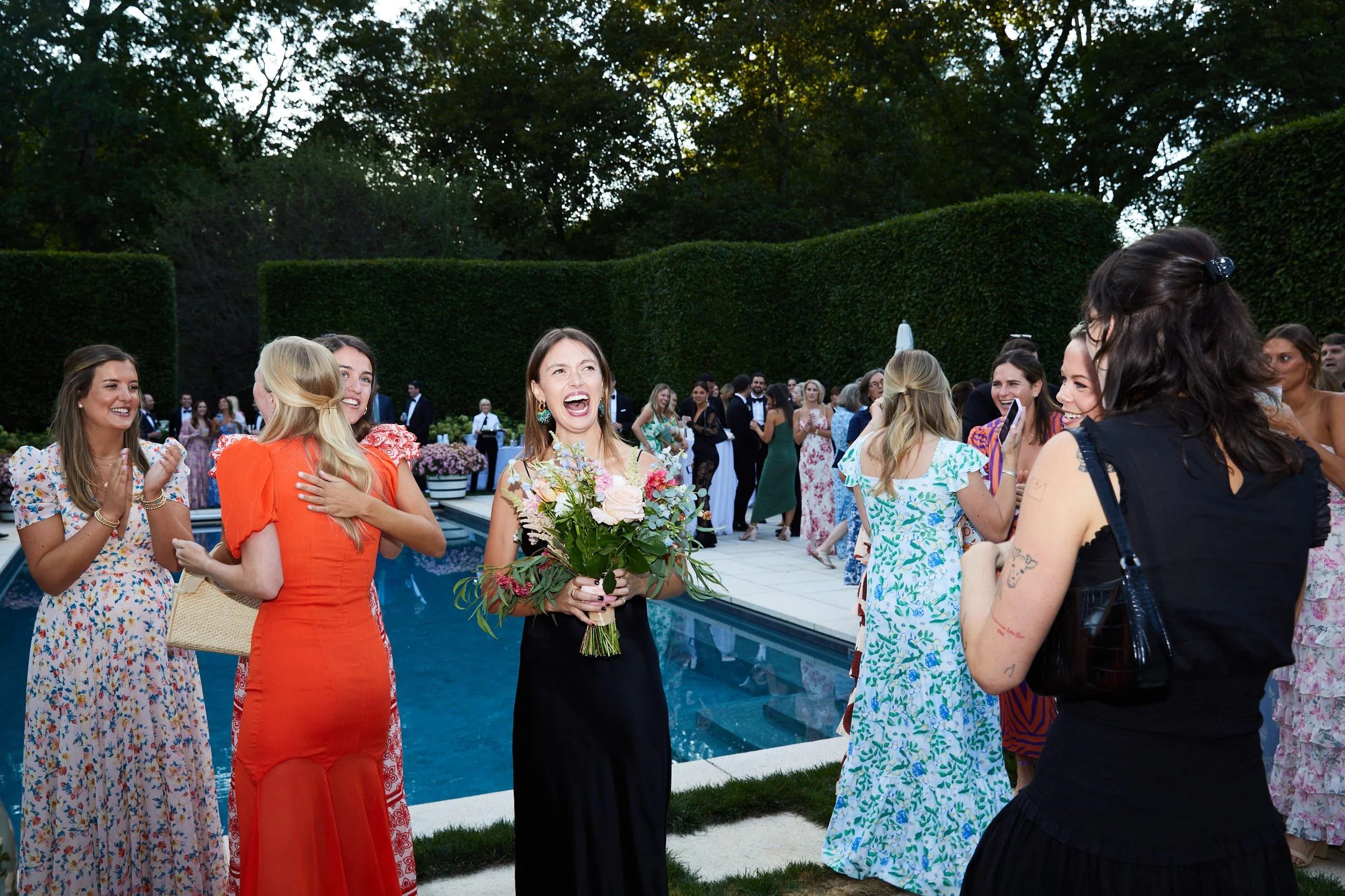 Group of women celebrating by a pool at an outdoor event, with one woman in a black dress holding a bouquet and smiling. Others are clapping, hugging, and smiling, with some dressed in floral dresses. The scene is lively with many people in the backg