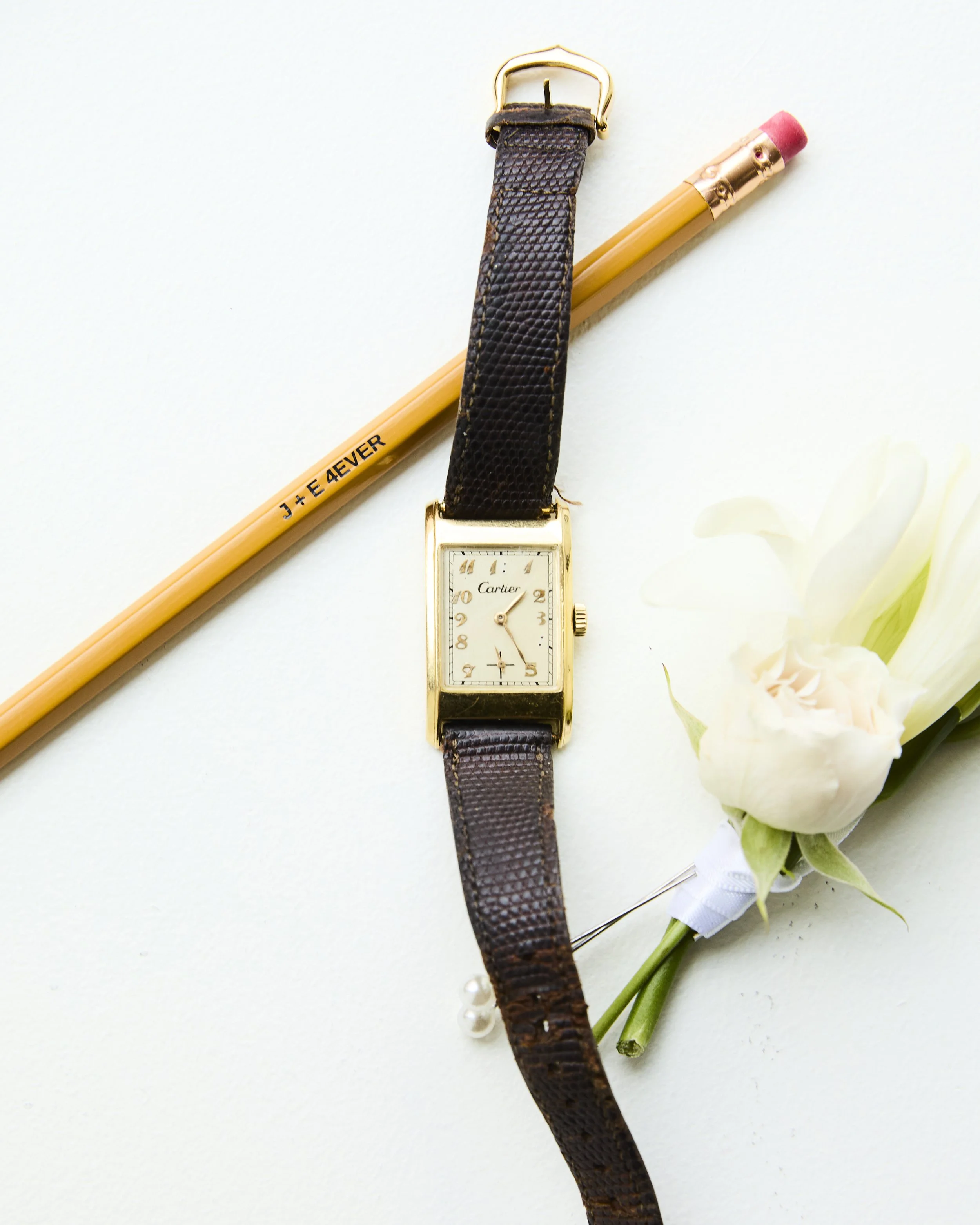 A Cartier wristwatch with a rectangular face and brown leather strap, placed on a white surface. Next to it is a light-colored wooden pencil and a white flower with a green stem.