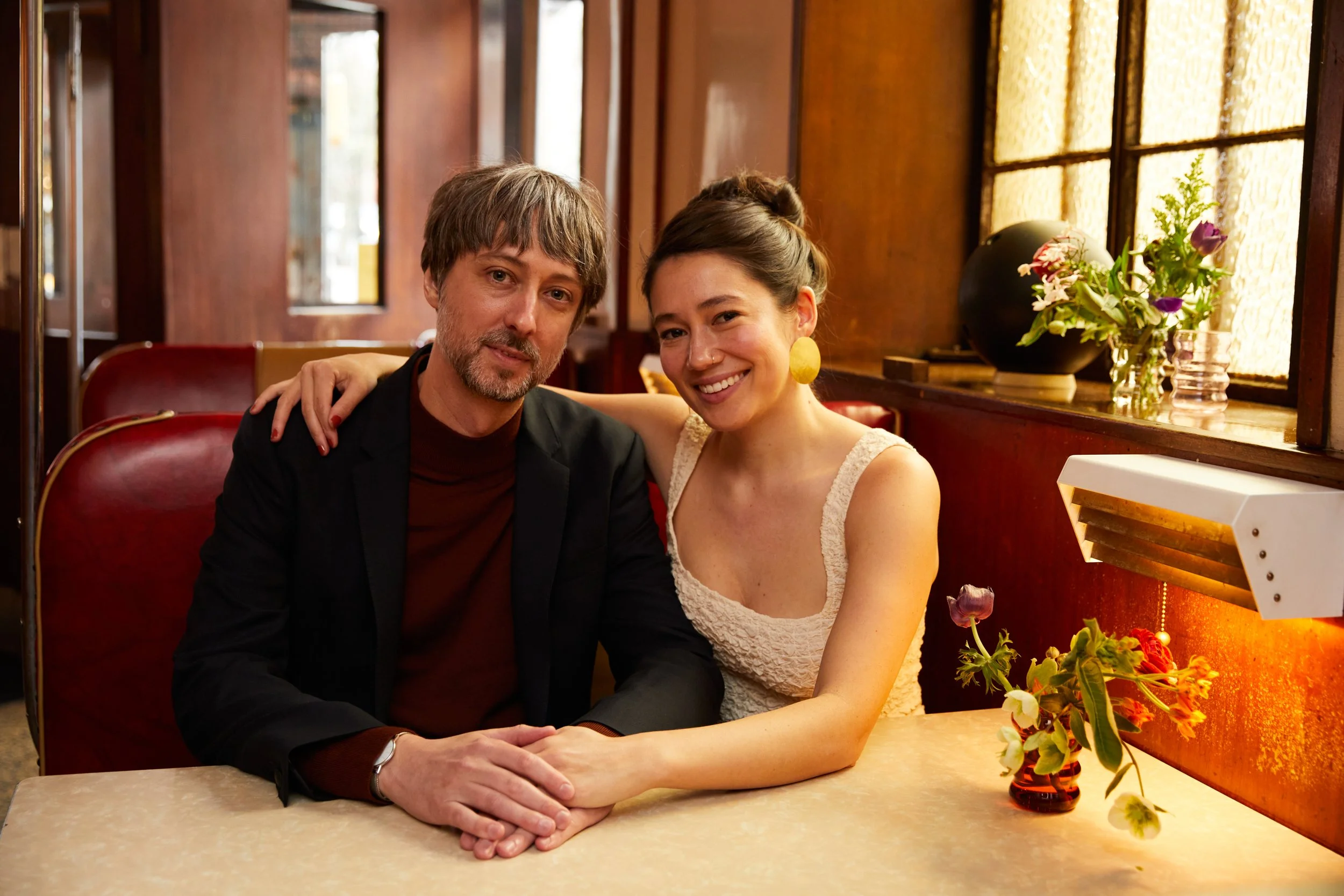 A couple sitting at a restaurant table, holding hands, smiling, with the woman having her arm around the man's shoulder.