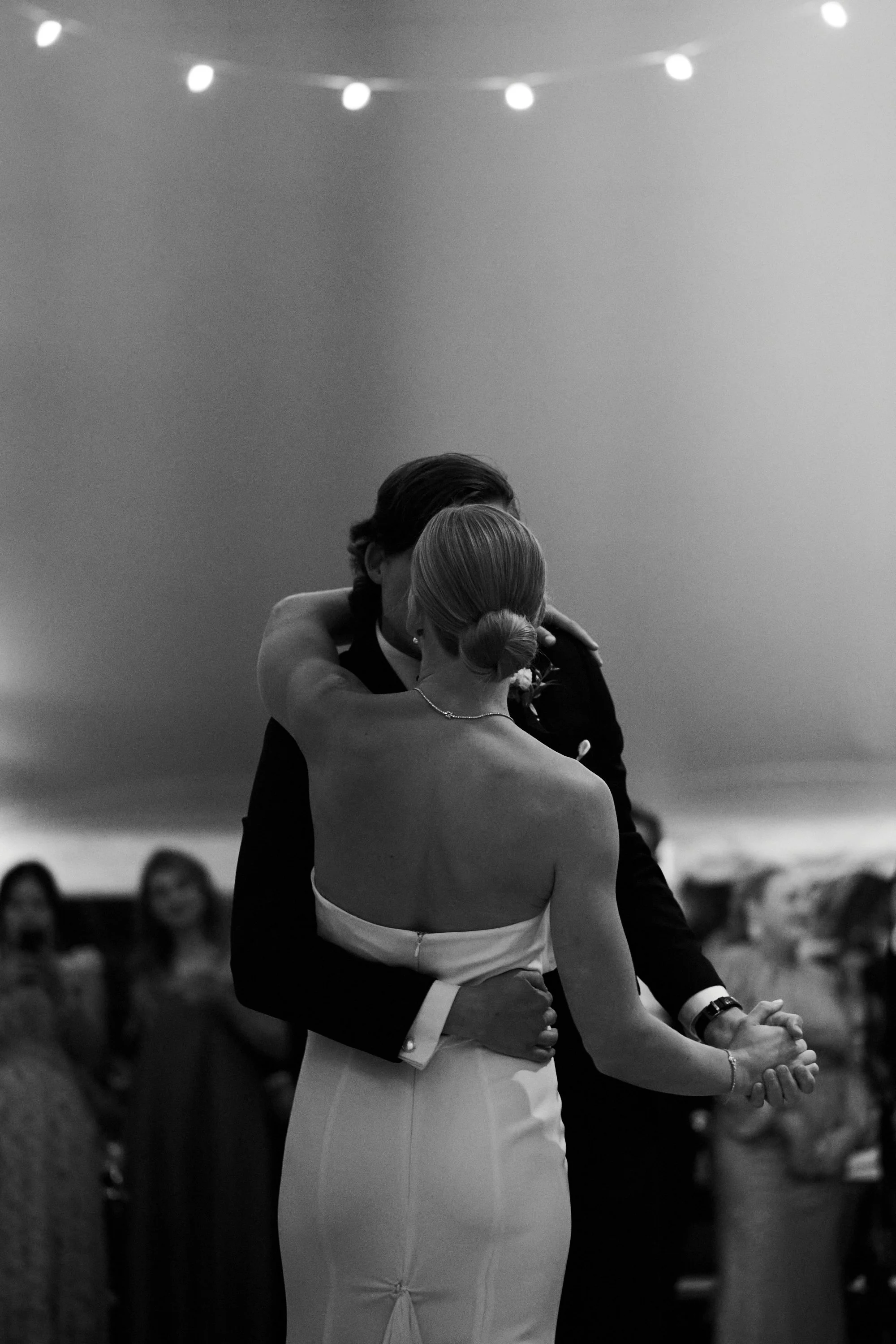 A black-and-white photo of a couple dancing closely at their wedding reception, with strings of lights overhead and guests in the background.