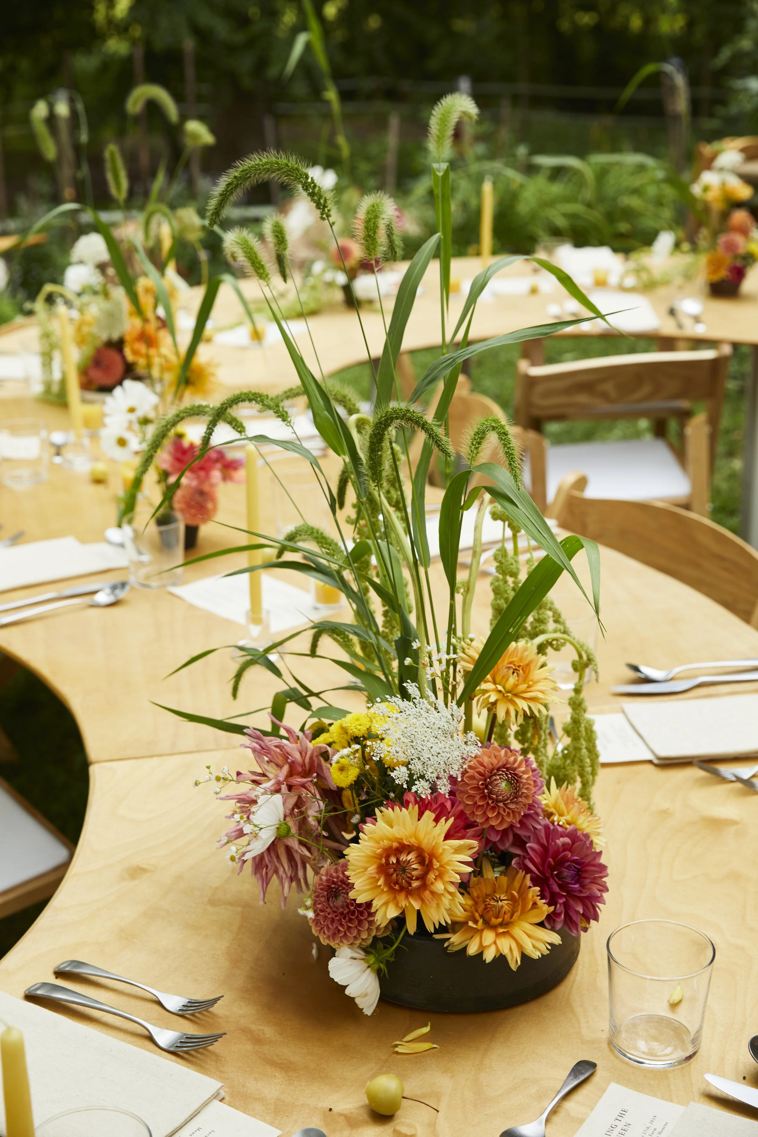 A floral arrangement with various colorful flowers and greenery as a centerpiece on a wooden dining table, set for an outdoor event in a green garden.