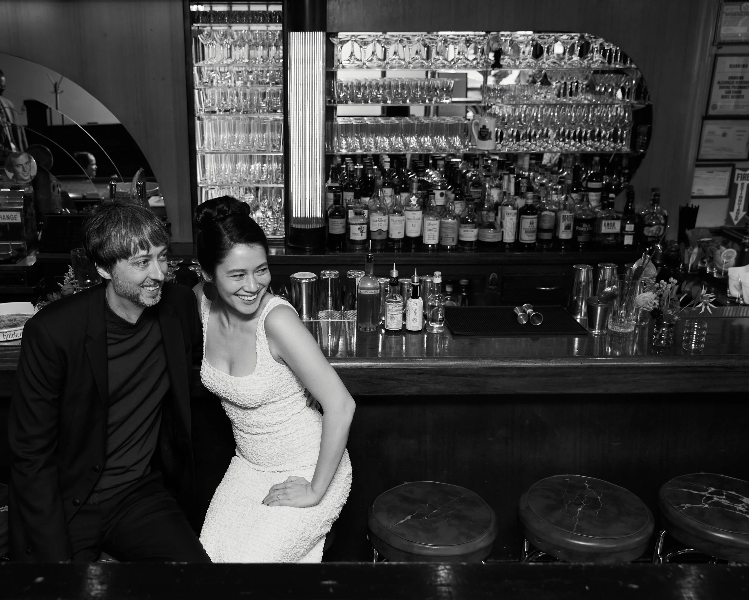 Two people, a man and a woman, sitting at a bar counter laughing. The bar area behind them is stocked with glasses, bottles, and bar tools.