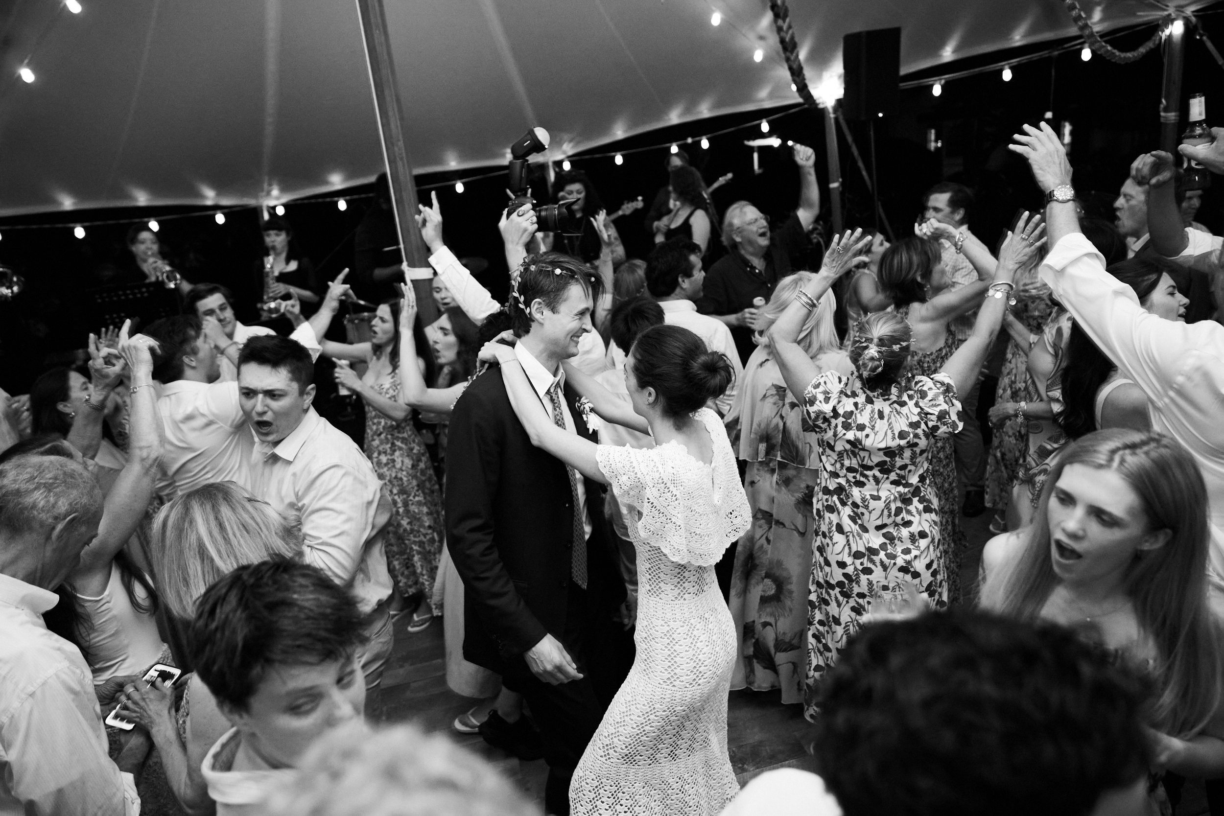 People dance and celebrate at a wedding reception under string lights, with the bride and groom in the center.