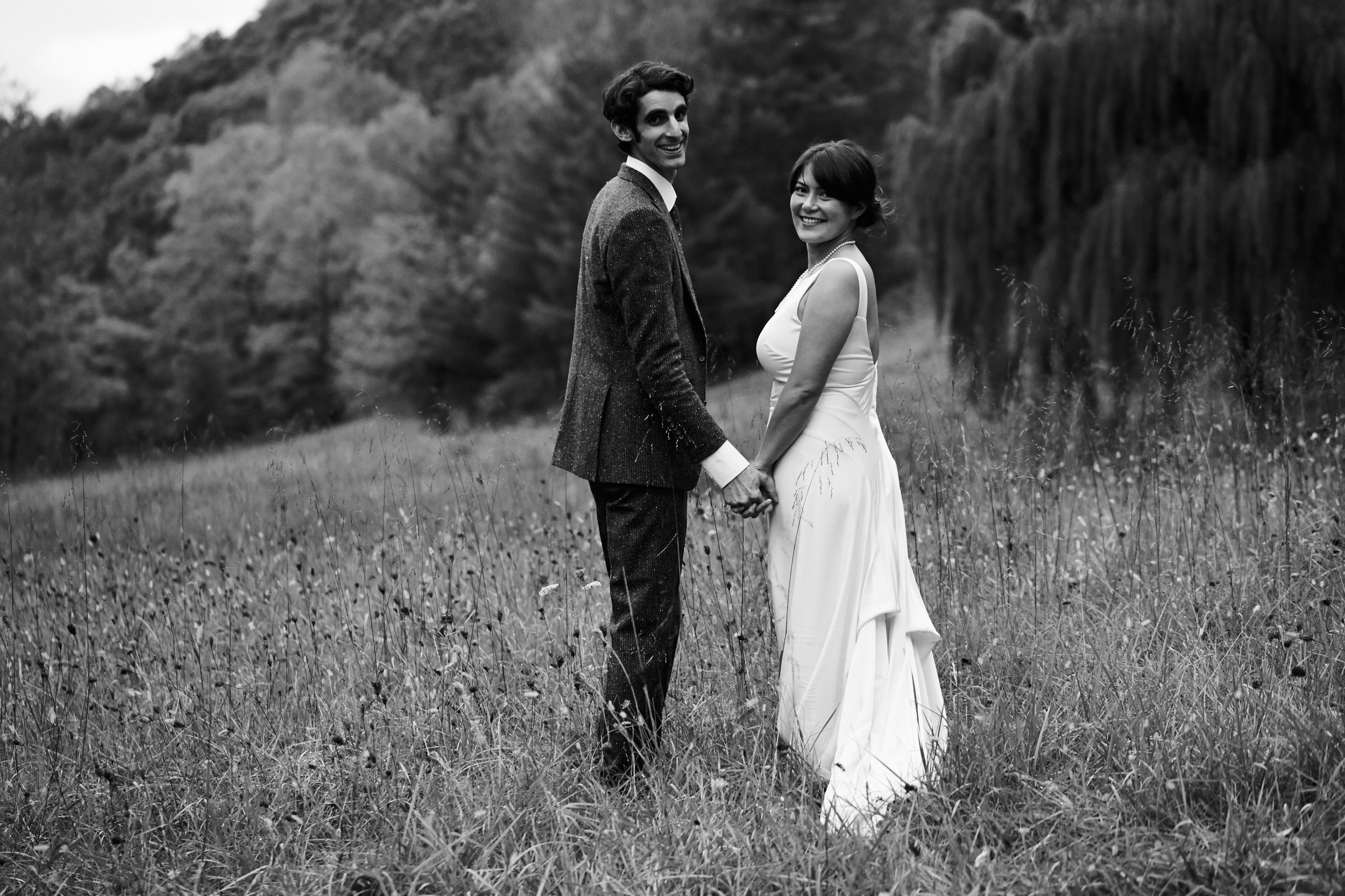 A black-and-white photo of a smiling man and woman holding hands in a grassy outdoor area, with trees in the background, dressed as a bride and groom.