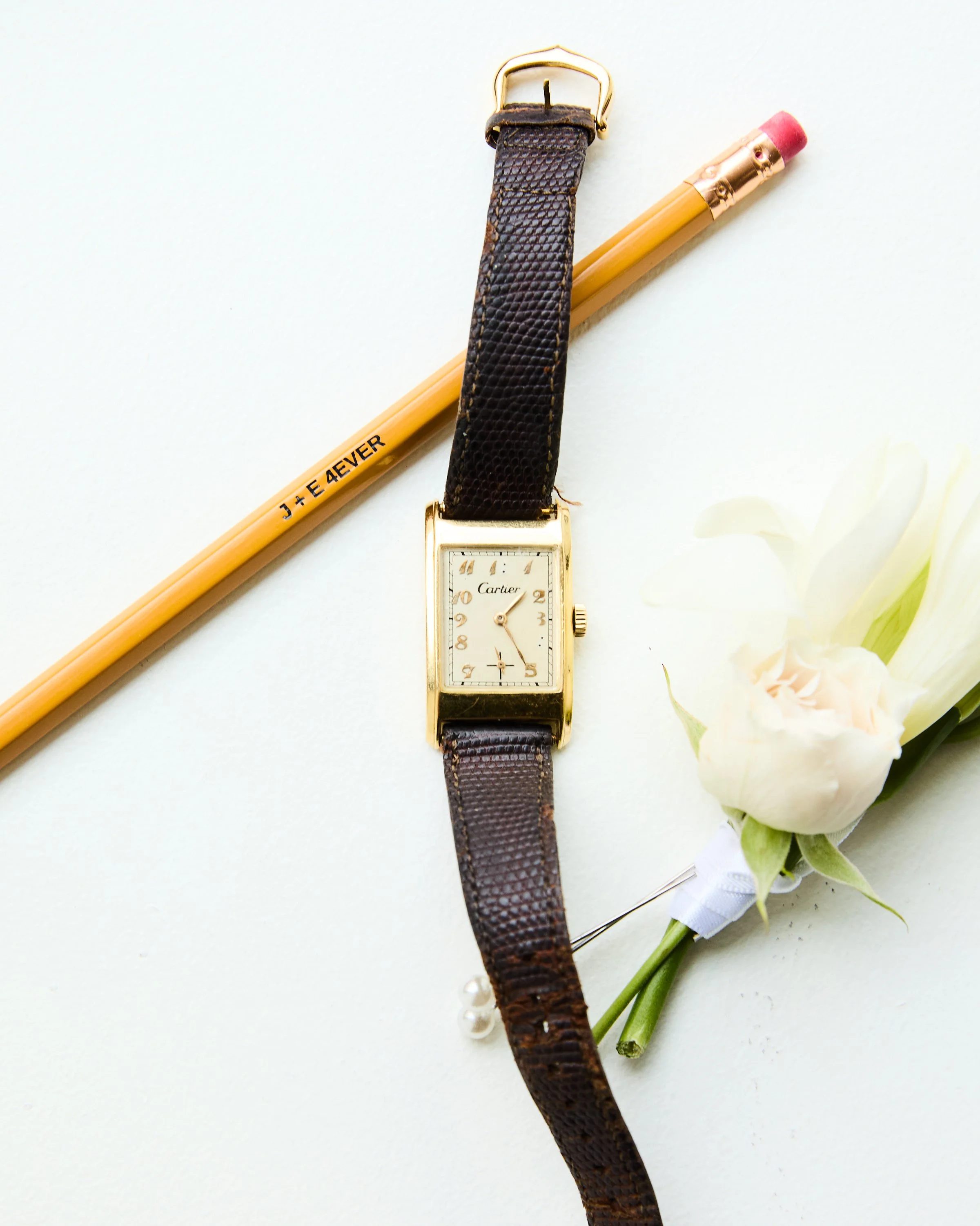 A gold Cartier wristwatch with a black leather strap, a pink eraser on a yellow pencil, and a white flower on a white background.