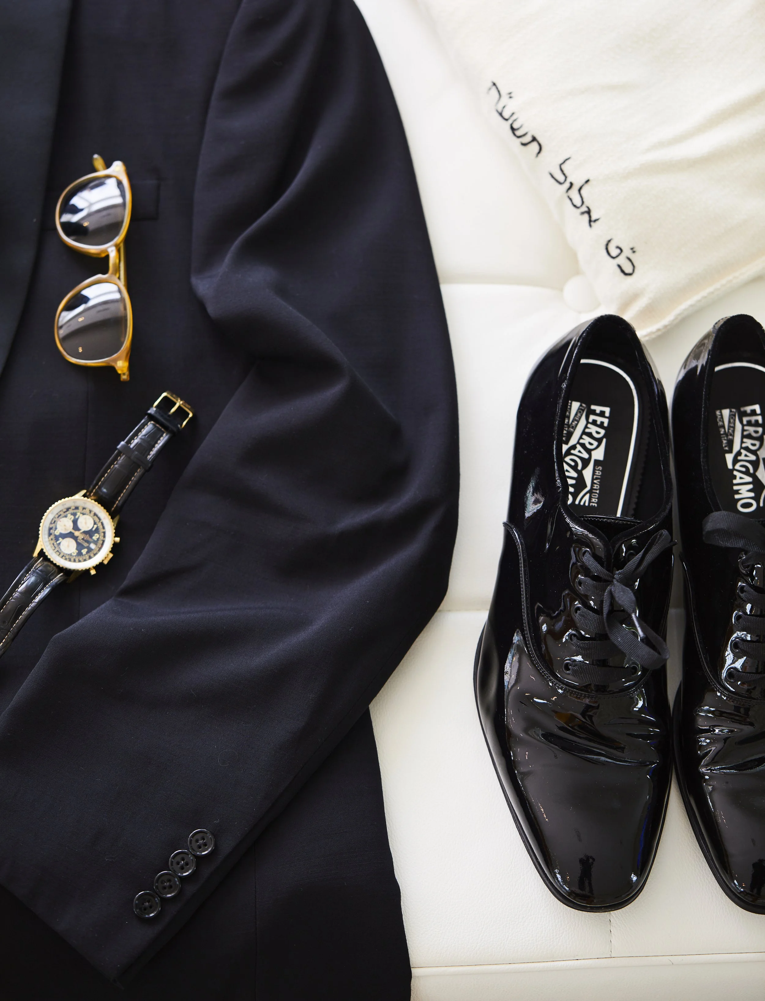 A black blazer with a gold-rimmed pair of sunglasses, a black leather strap watch, and shiny black leather shoes on a white surface with a pillow that has black embroidery.