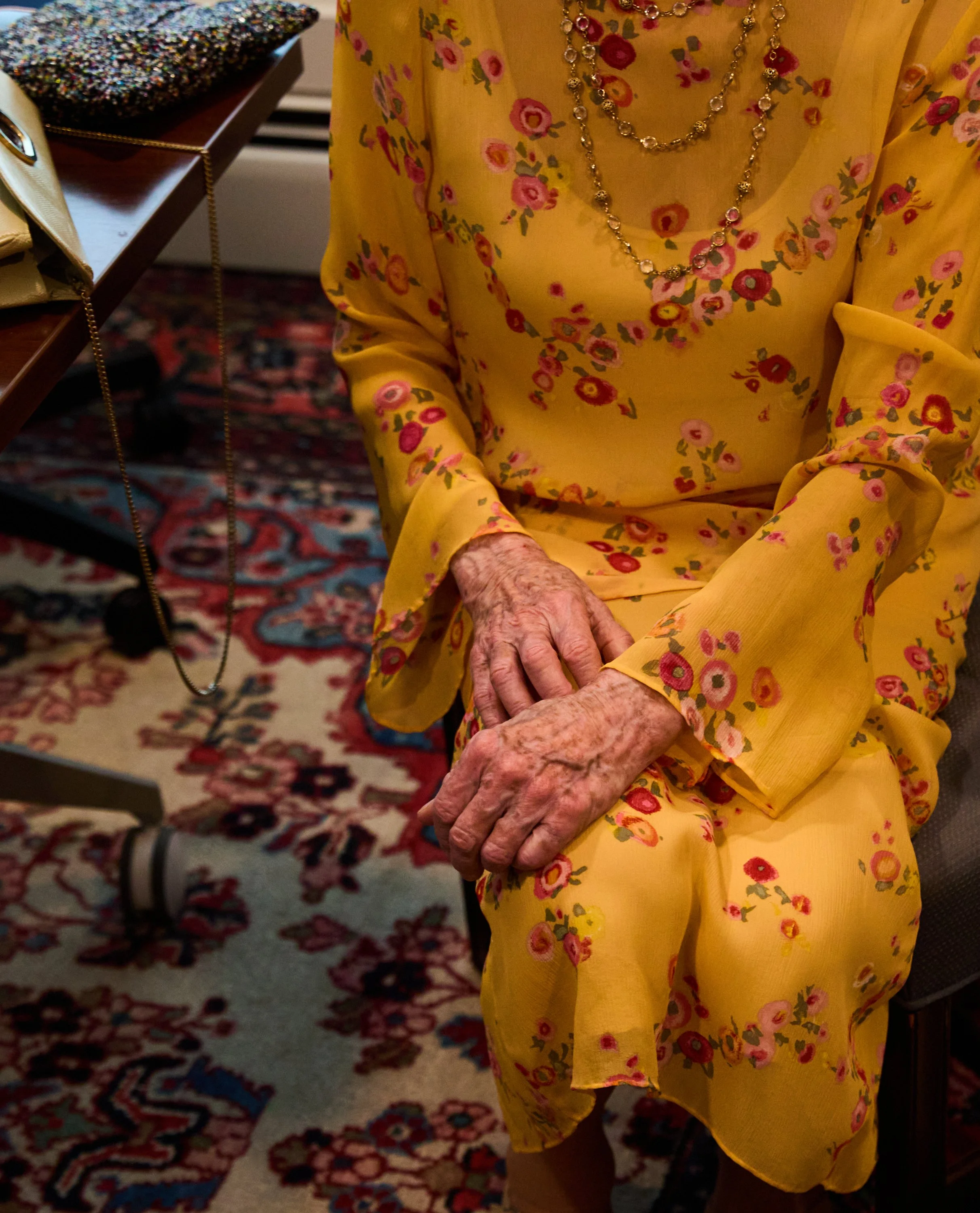Close-up of an elderly woman's hands resting on her lap, wearing a yellow floral dress and layered necklaces, seated on a patterned carpet with a piece of furniture and accessories nearby.