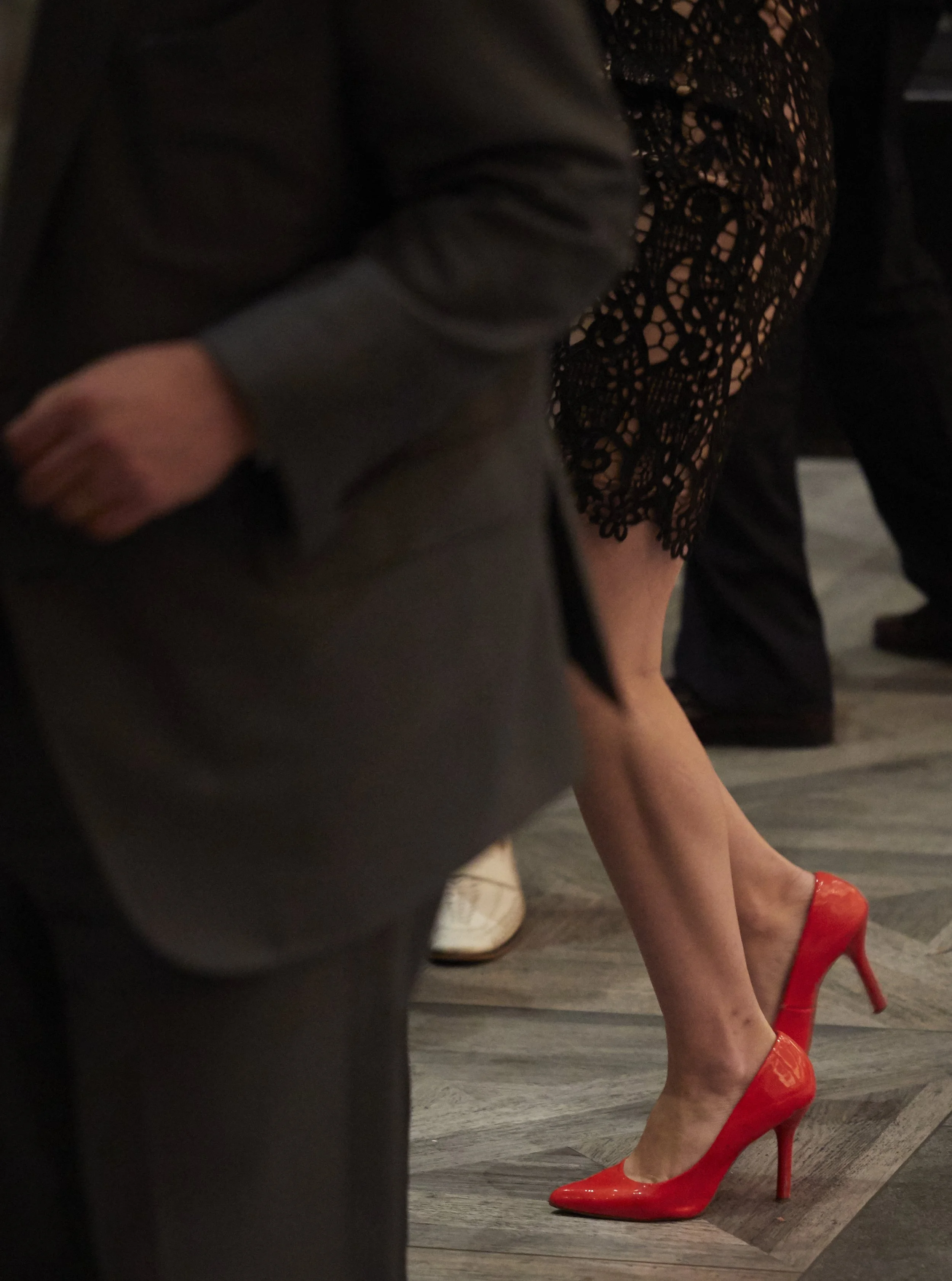 A woman wearing red high heels and a black lace dress standing on a wooden floor. Part of a person in a dark suit is visible in the foreground.