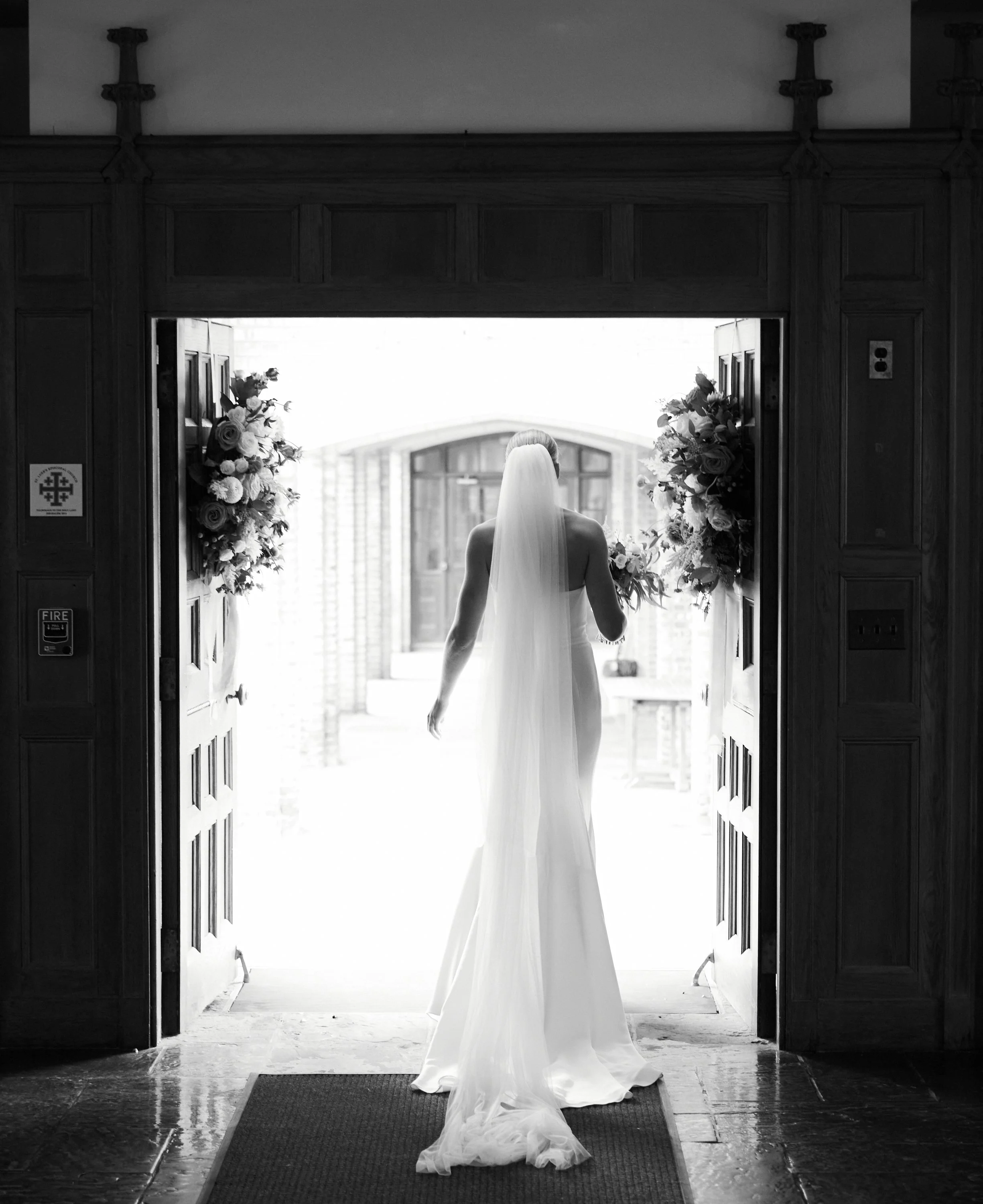 Silhouette of a bride in a wedding dress with a veil, holding a bouquet, standing in a doorway decorated with flowers, looking outside.