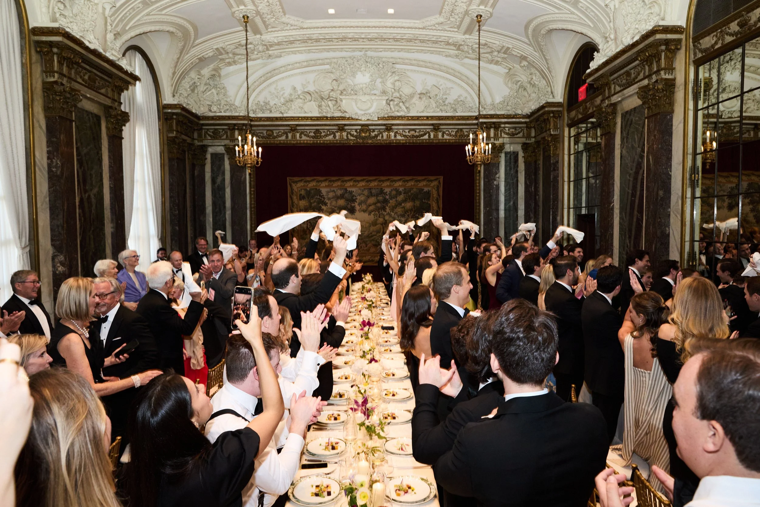 People dressed in formal attire at a banquet or celebration, waving napkins in the air in a grand, ornately decorated room with chandeliers and mirrors.