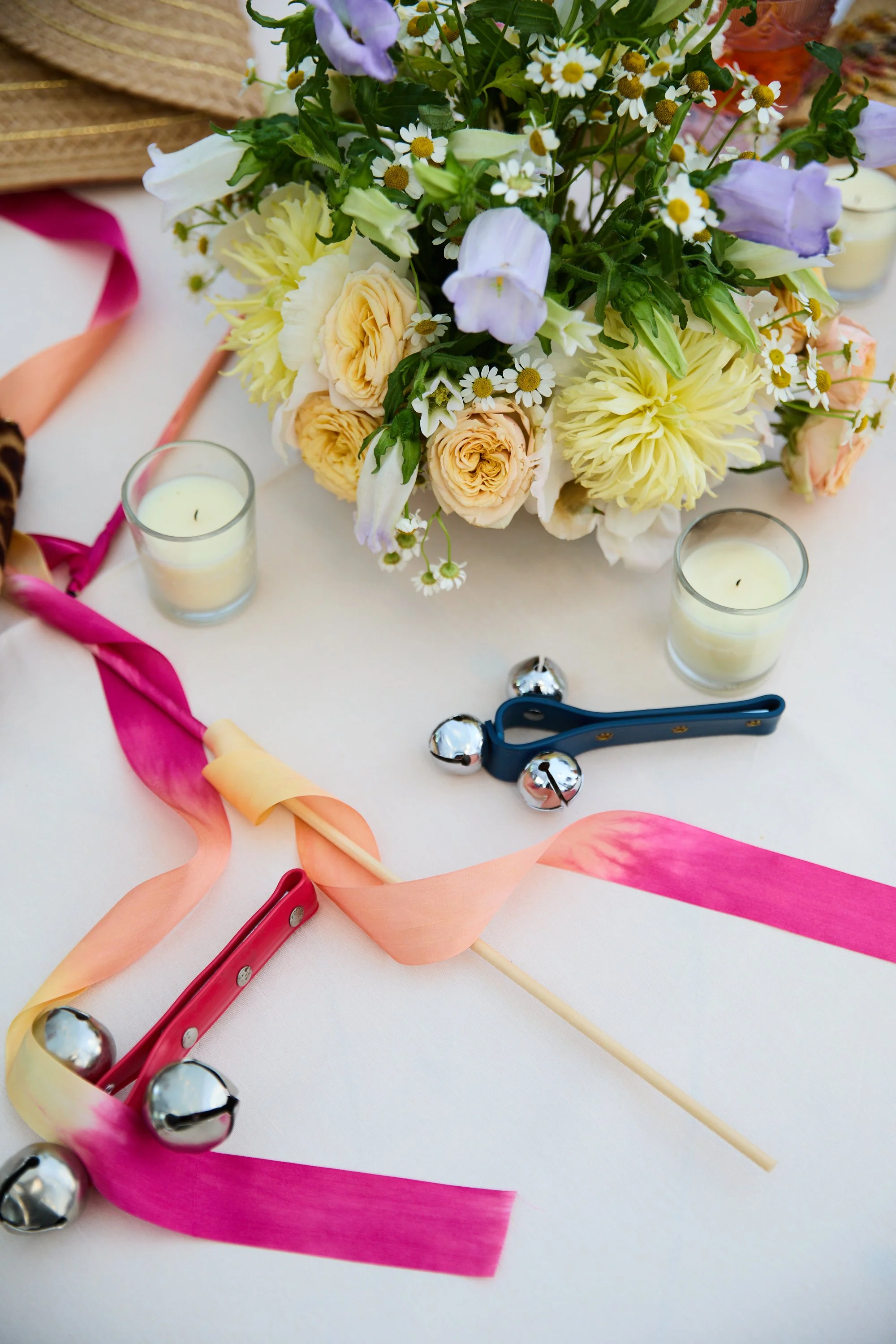 A floral centerpiece with white, yellow, and purple flowers on a table surrounded by candles and decorative metal bells, with colorful ribbons and small musical instruments.