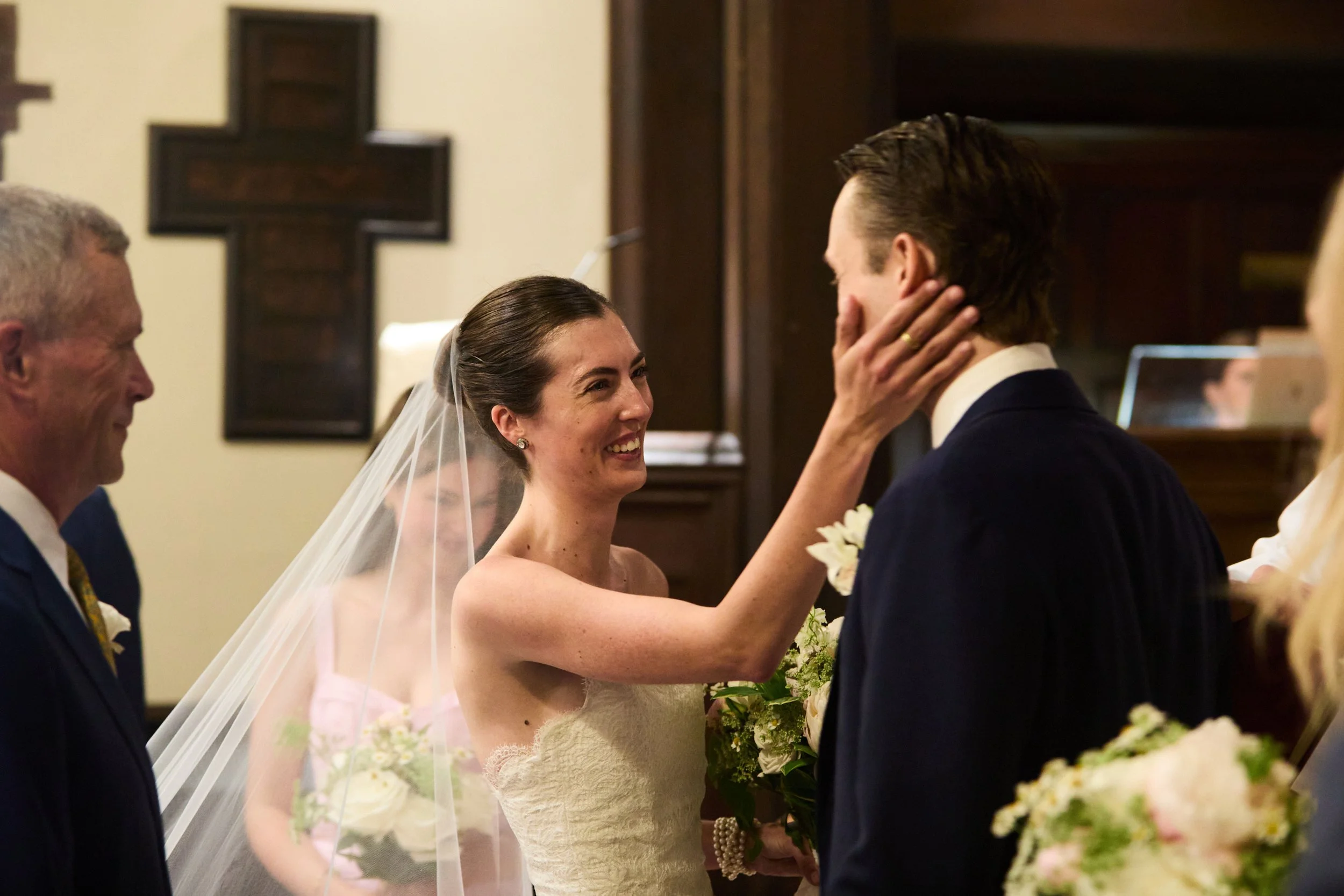 A bride smiling and holding a bouquet, touching a groom's face during a wedding ceremony, with a priest and another woman in the background.