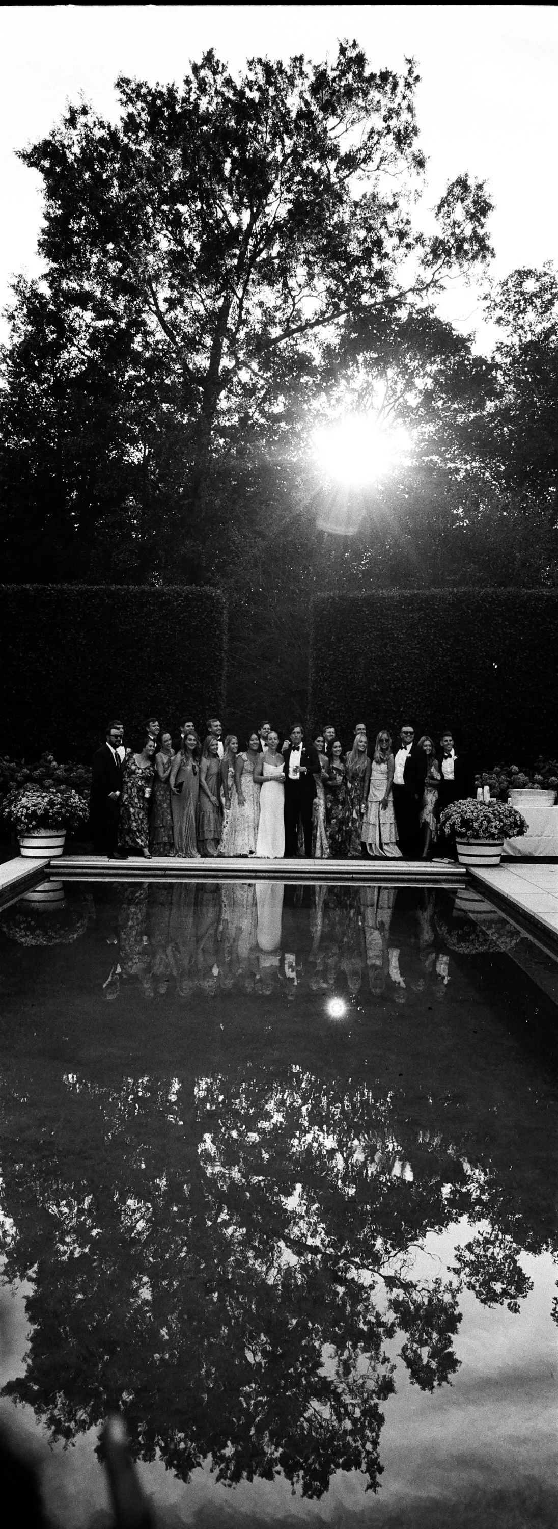 A large group of people dressed in formal attire standing together by a poolside in a garden setting during the evening, with trees and the sunset visible in the background, and their reflection in the pool water.