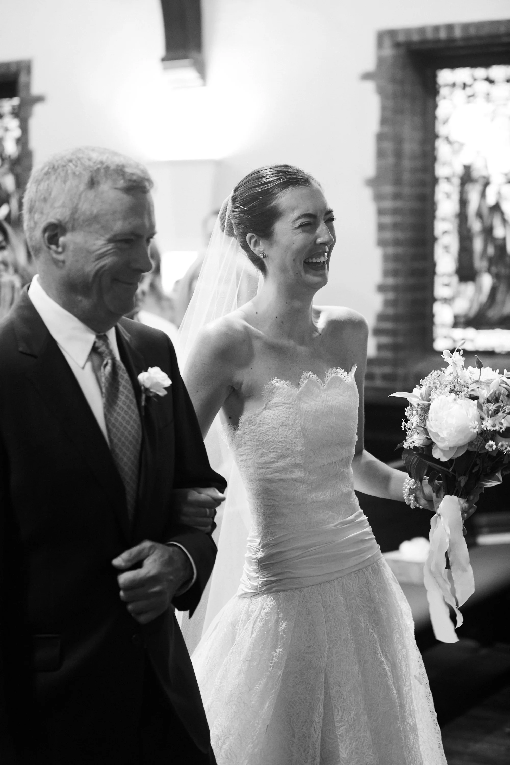 A bride smiling and holding a bouquet walking down the aisle with an older man in a suit, inside a church.