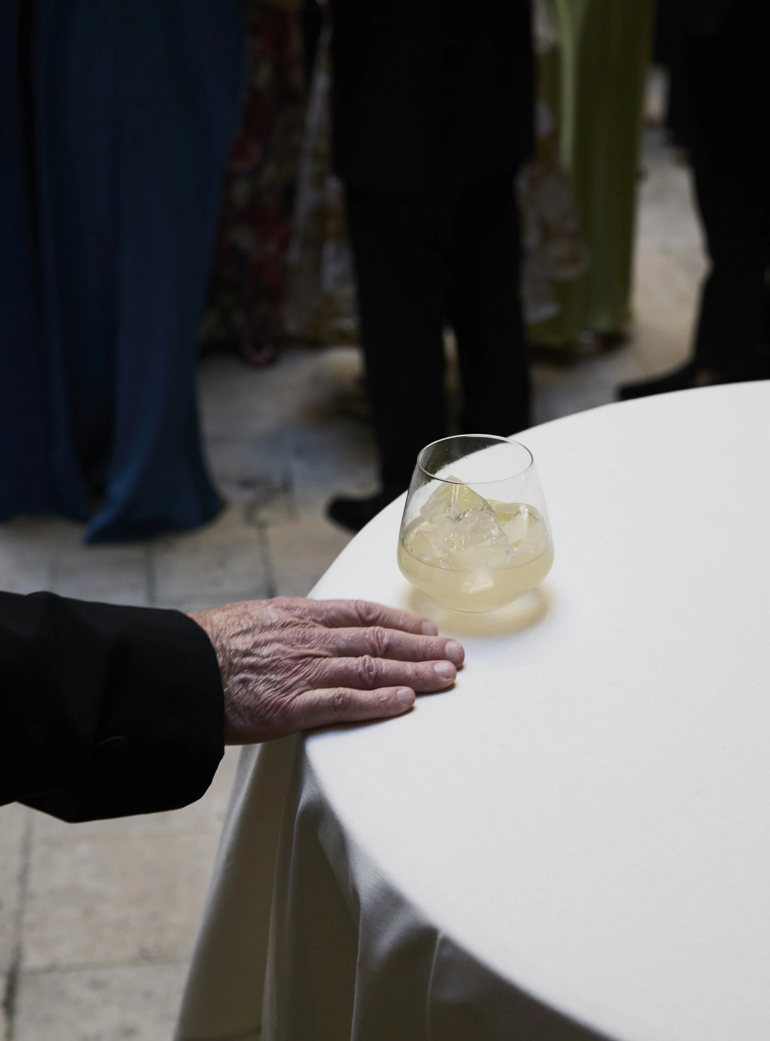 A hand resting on a white table next to a glass of light-colored beverage with ice cubes, with people standing in the background.