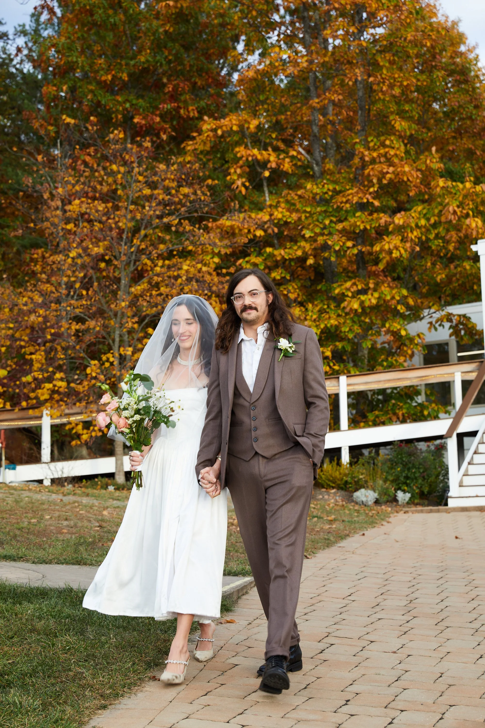A bride and groom holding hands walking outdoors, with colorful fall foliage in the background. The bride is wearing a white dress and veil, holding a bouquet of flowers. The groom is dressed in a brown suit with a white shirt and boutonniere.