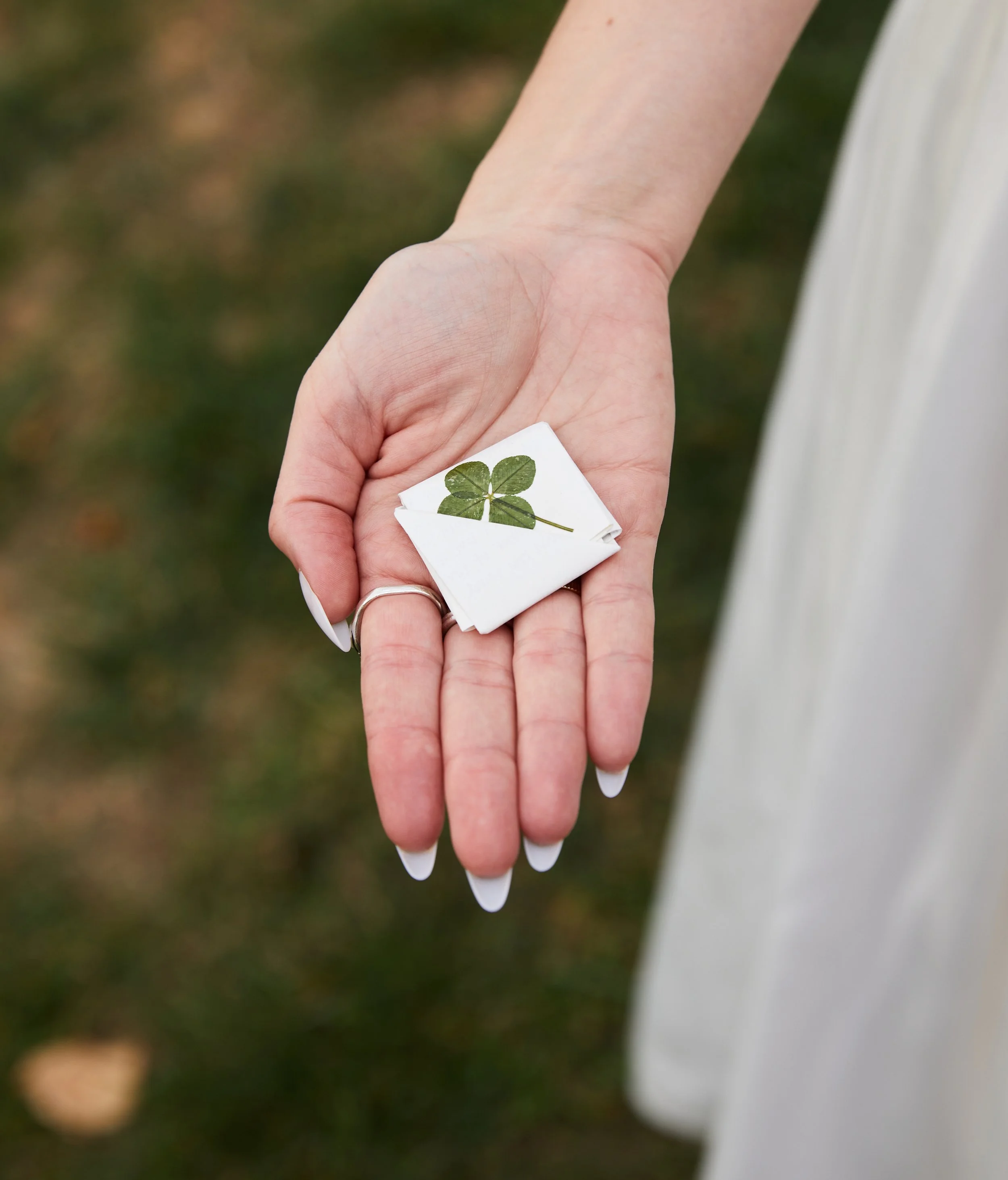 A hand holding a small folded napkin with a four-leaf clover printed on it, outdoors with grassy ground in the background.