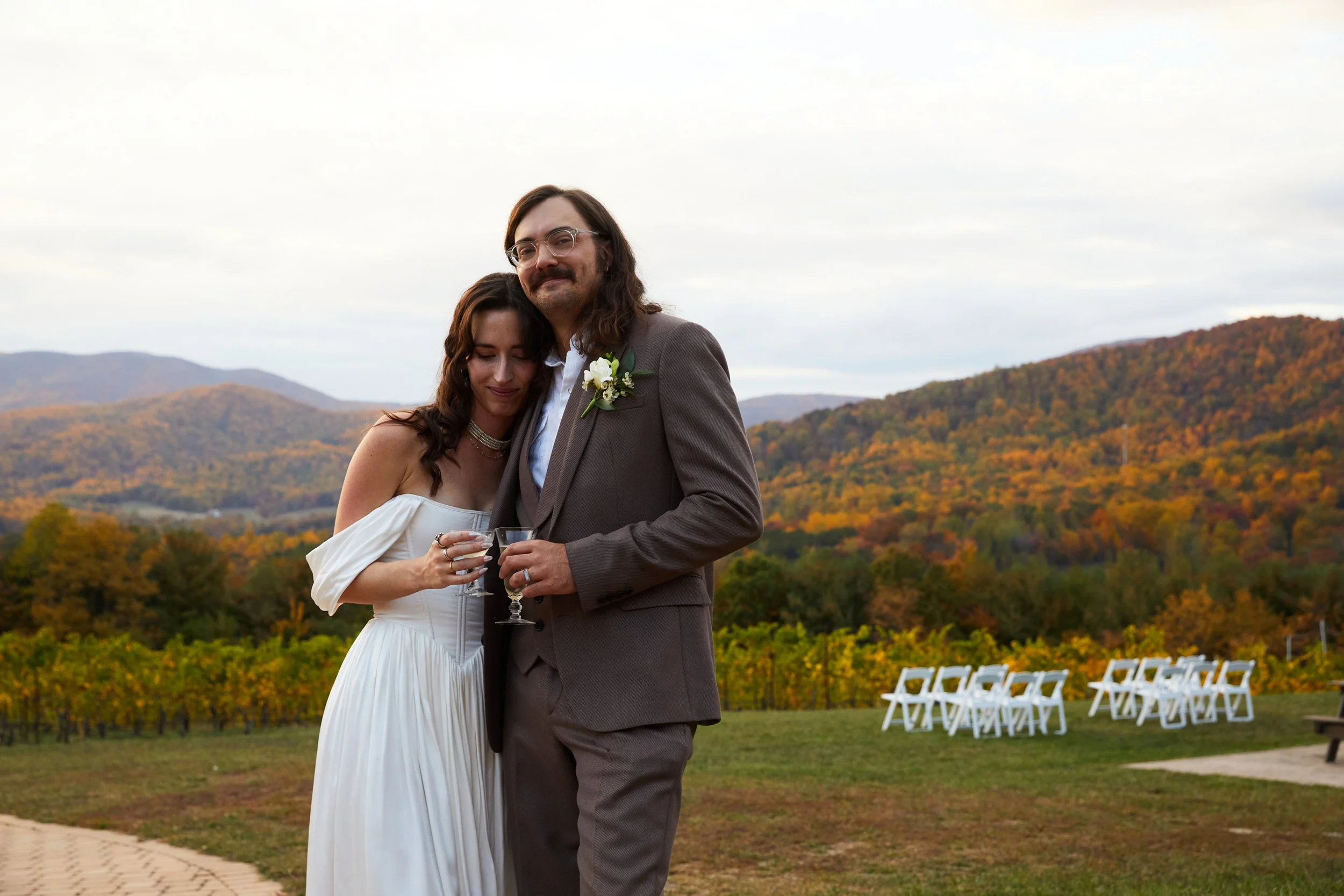 A newly married couple with the woman in a white dress and the man in a gray suit, holding glasses, standing outdoors with mountains and autumn foliage in the background during daytime.