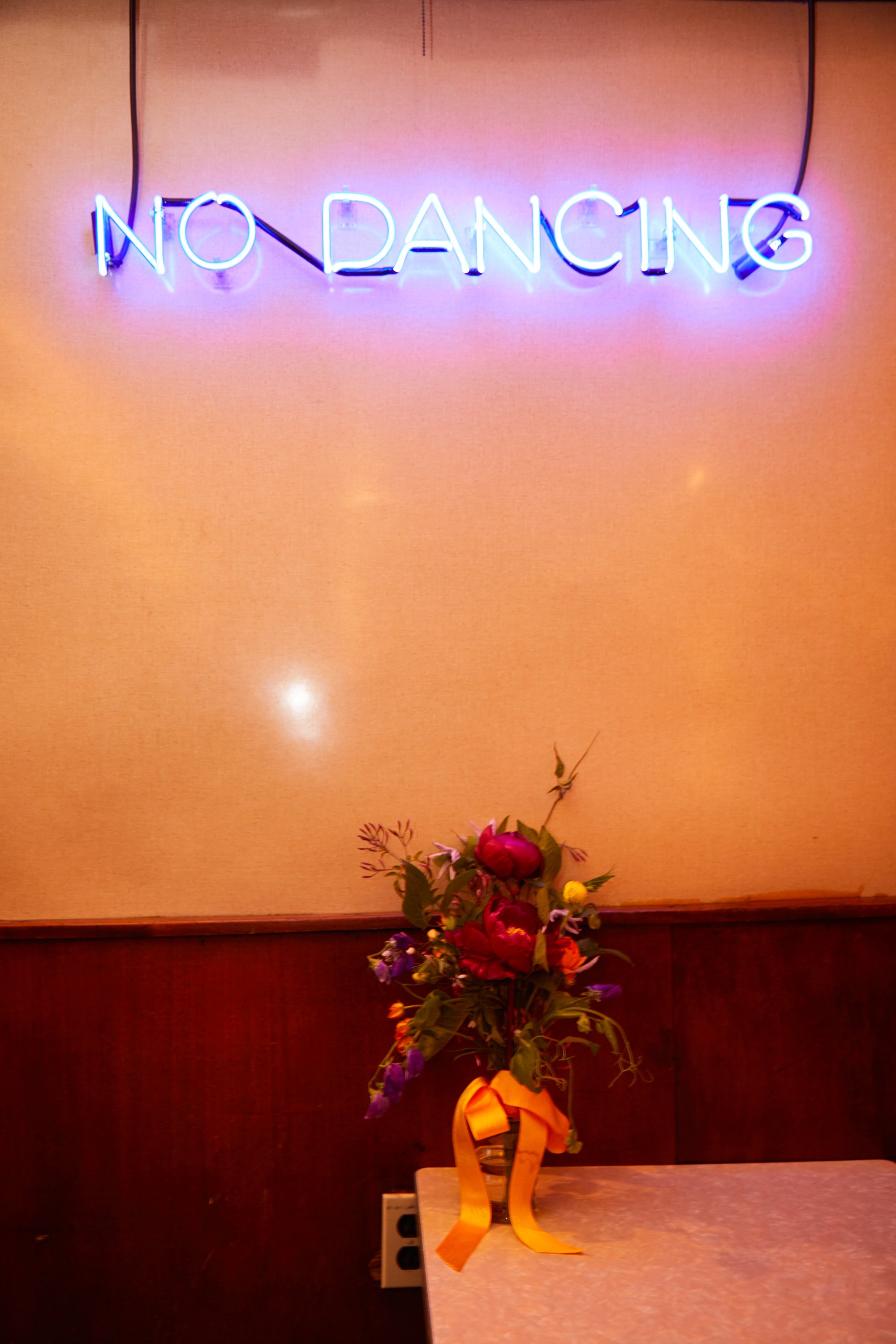 A neon sign on a pink wall that says 'NO DANCING.' Below the sign, there is a table with a flower arrangement wrapped with a yellow ribbon.