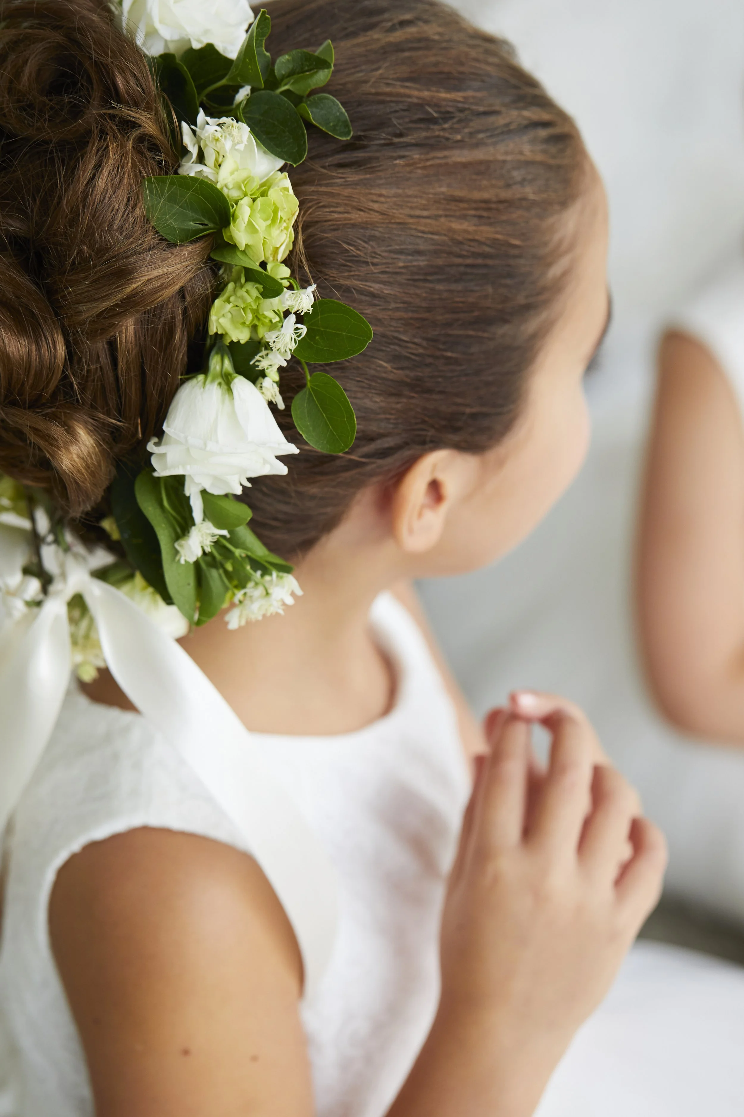 A young girl with brown hair styled in an elegant updo, decorated with white and green flowers and leaves.